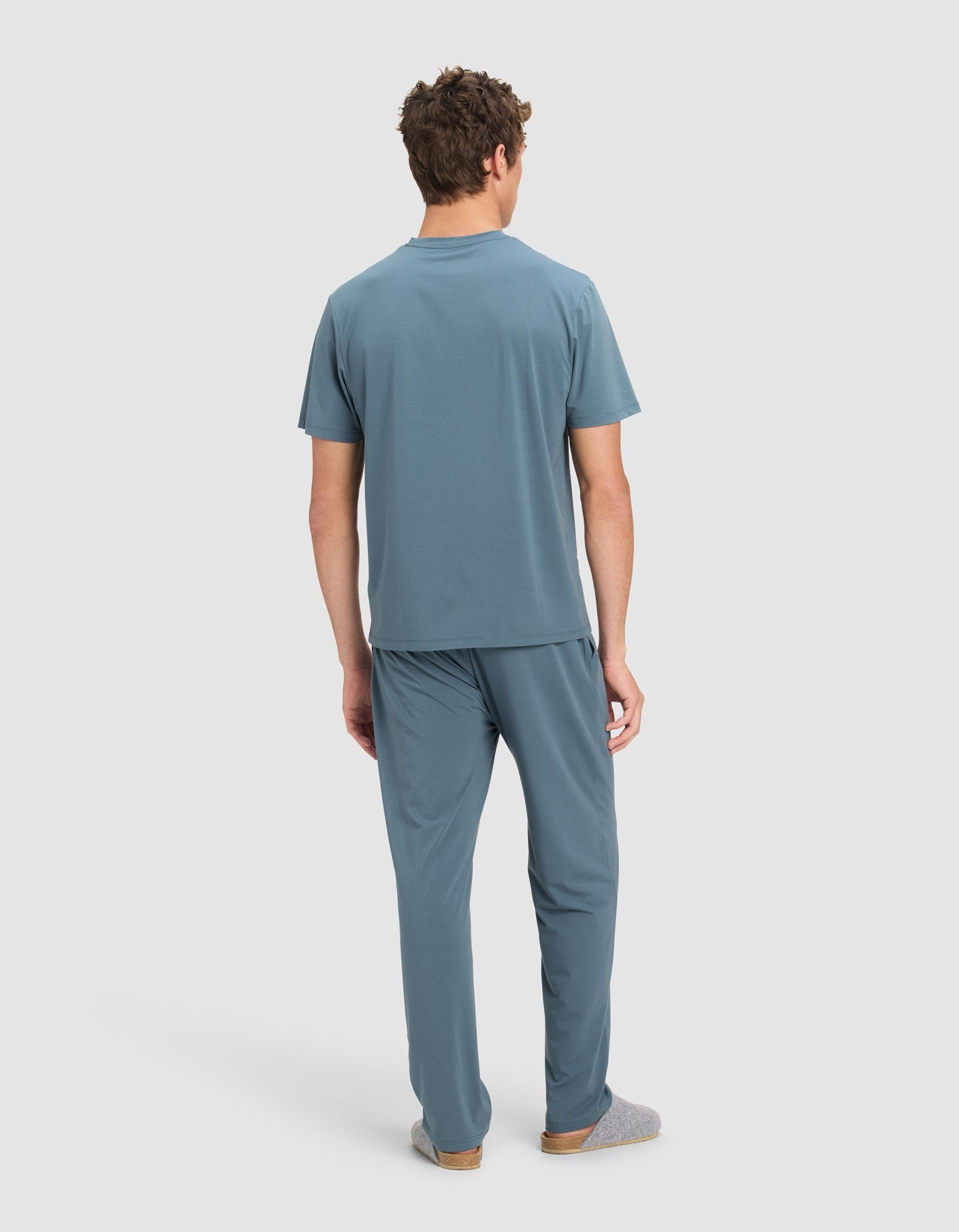 A man faces away, wearing the Cozy Earth Men's First Layer Pajama Short Sleeve Tee & Pant Set in blue with gray slippers, against a plain white background.