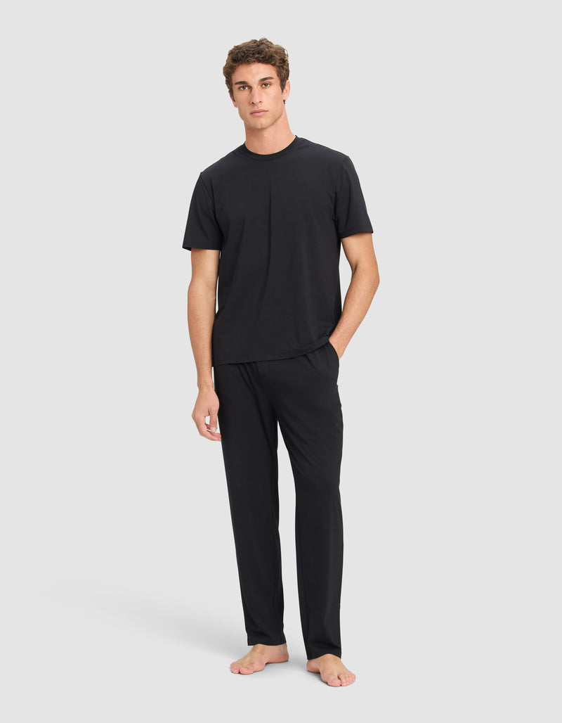 A man stands barefoot against a plain background, wearing the Cozy Earth Men's First Layer Pajama Short Sleeve Tee & Pant Set in black with his left hand in his pocket.