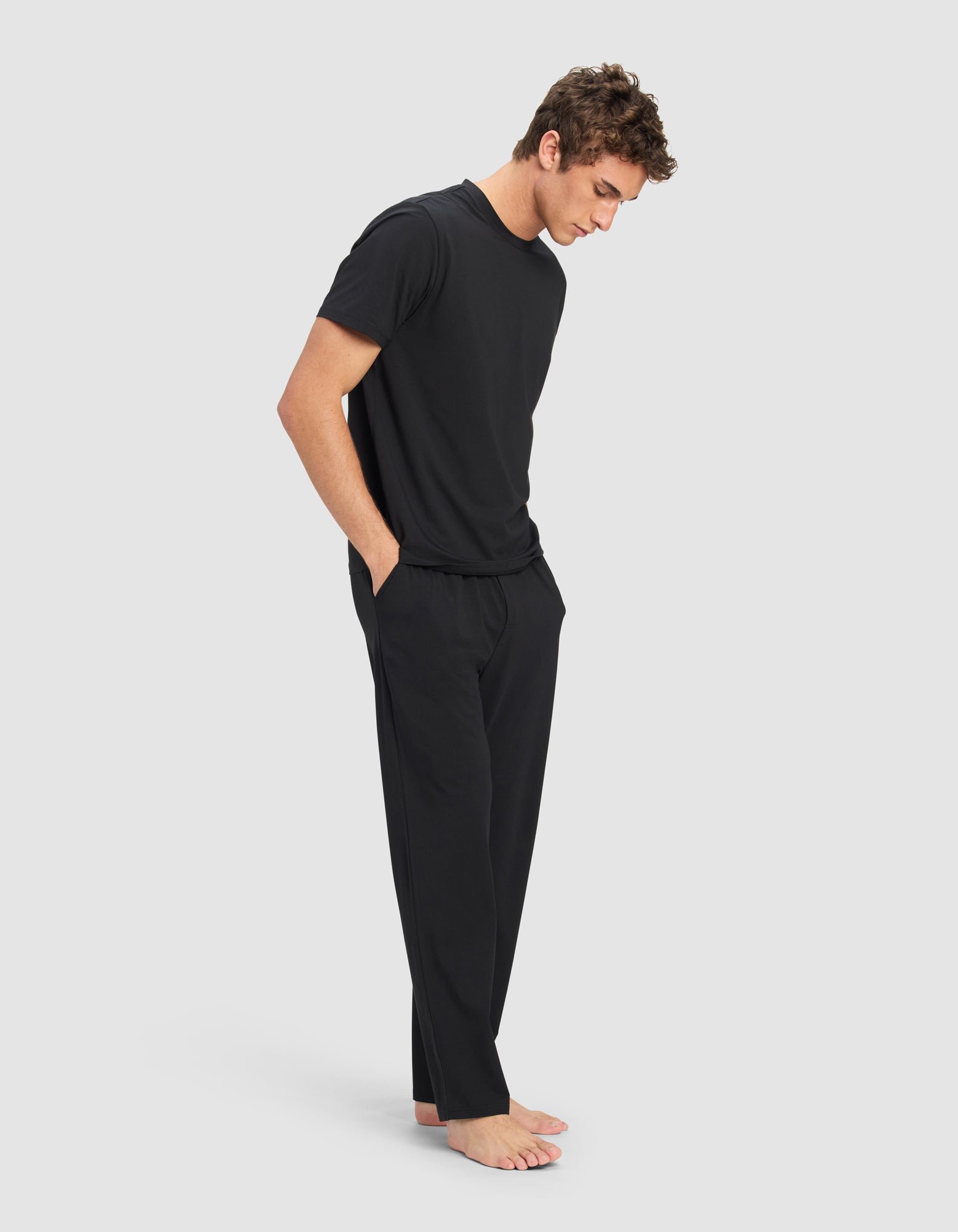 A man stands barefoot against a plain background, wearing the Cozy Earth Men's First Layer Pajama Short Sleeve Tee & Pant Set in black, with his hands in his pockets and looking down.|Color:Jet Black