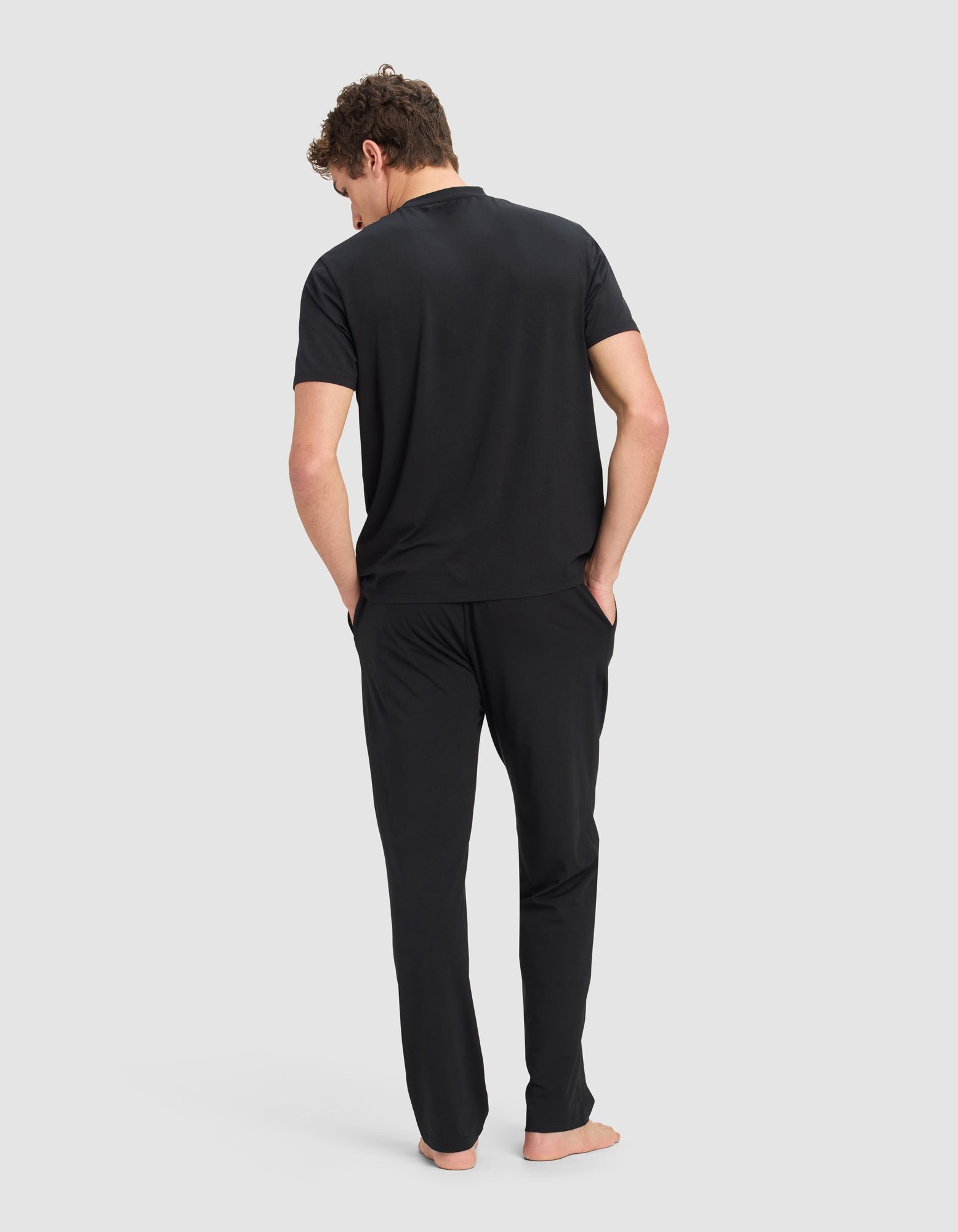 A man stands barefoot with his hands in his pockets, wearing the Cozy Earth Men's First Layer Pajama Short Sleeve Tee & Pant Set in black, facing away against a plain light gray background. His head is slightly tilted downward.|Color:Jet Black