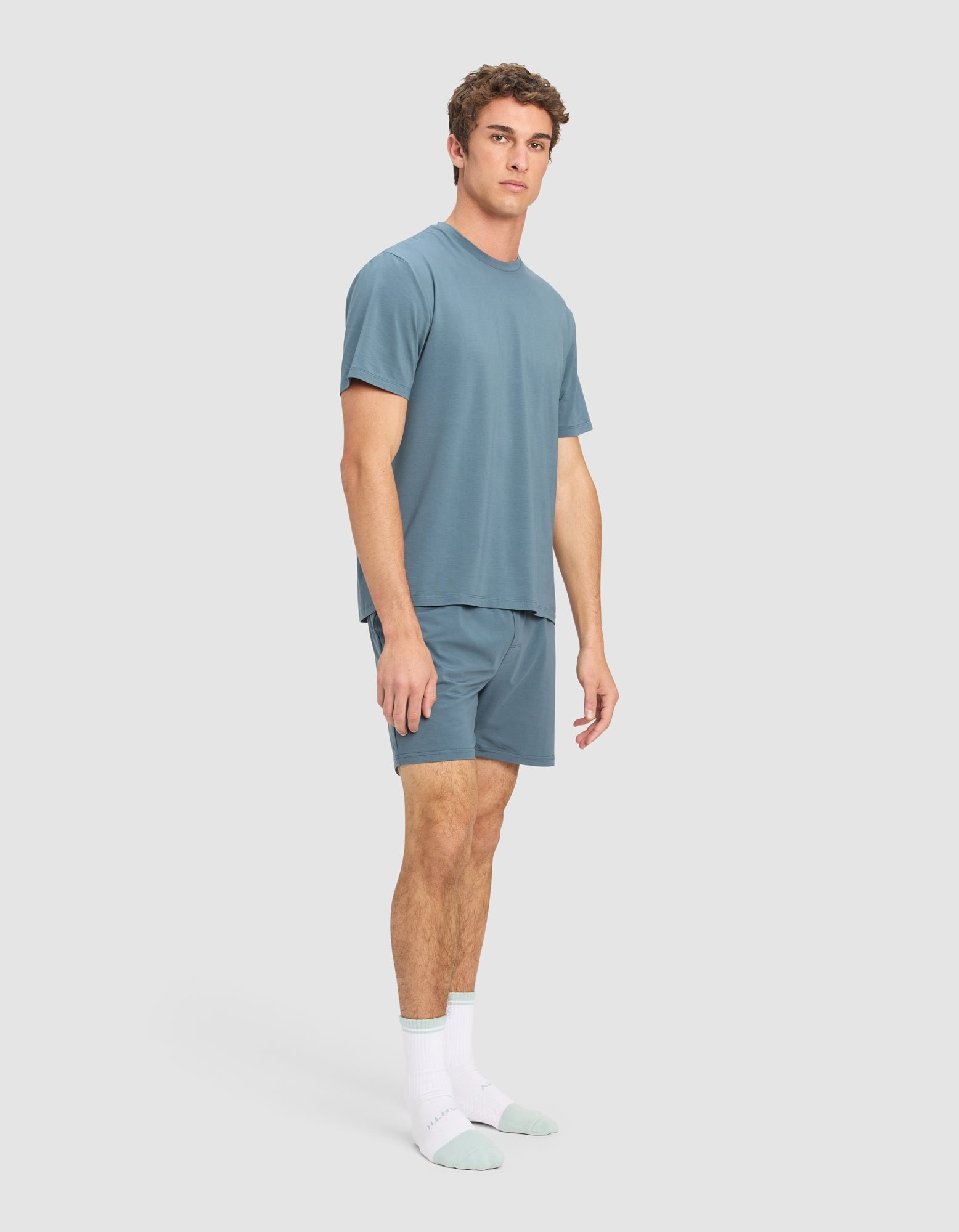 A man stands in a relaxed pose wearing the Cozy Earth Men's First Layer Pajama Short Sleeve Tee & Short Set and white socks with light green toes and heels, against a plain light gray background.|Color:Dawn