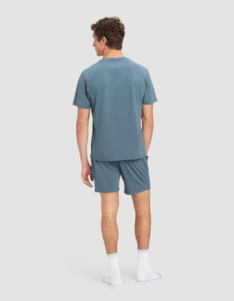 A man faces away, wearing the Men's First Layer Pajama Short Sleeve Tee & Short Set by Cozy Earth in blue with white socks featuring green accents. The background is plain light gray.