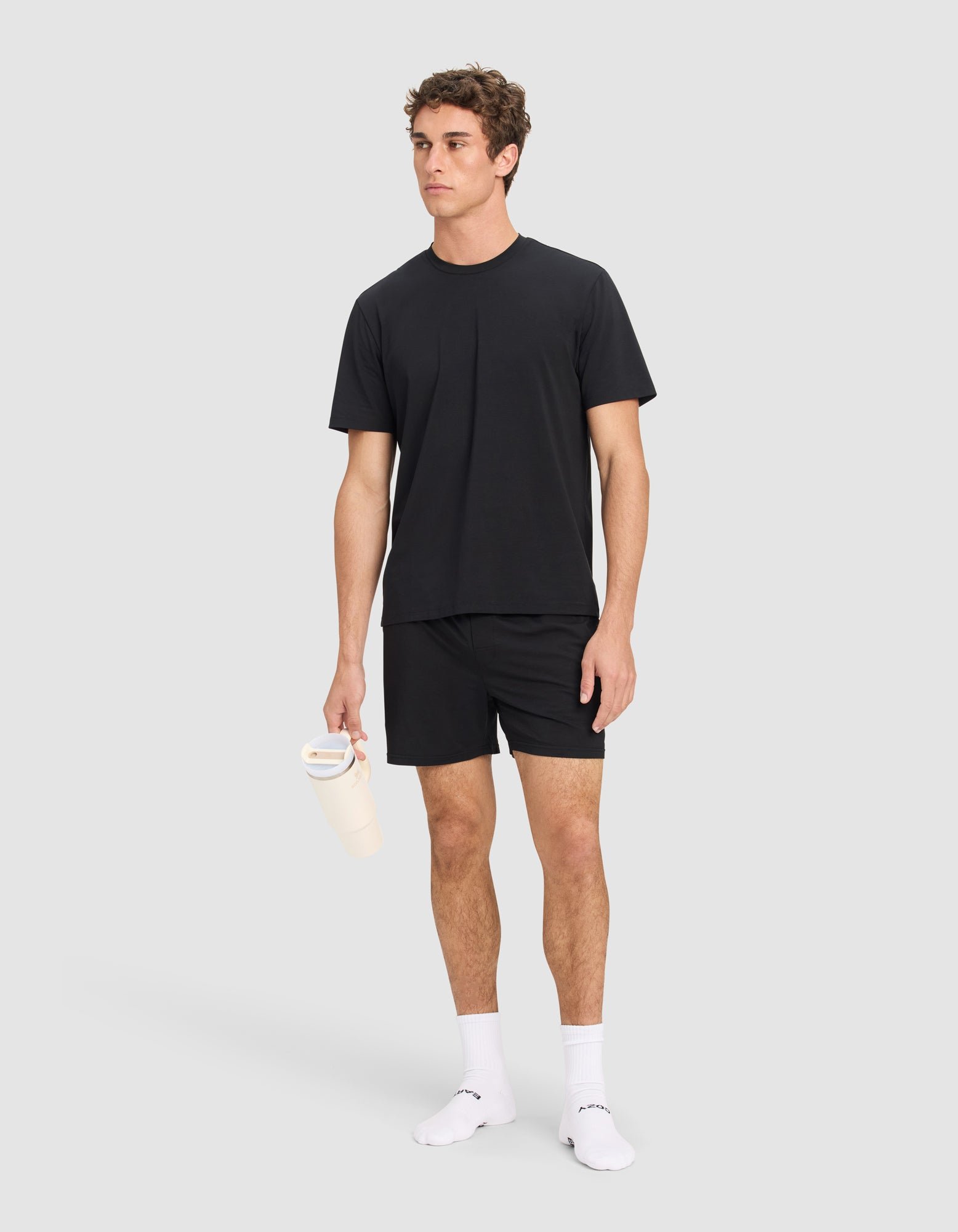 A man wears the Cozy Earth Men's First Layer Pajama Short Sleeve Tee & Short Set in black, paired with white socks and holding a light-colored water bottle, posing against a plain light gray background.|Color:Jet Black