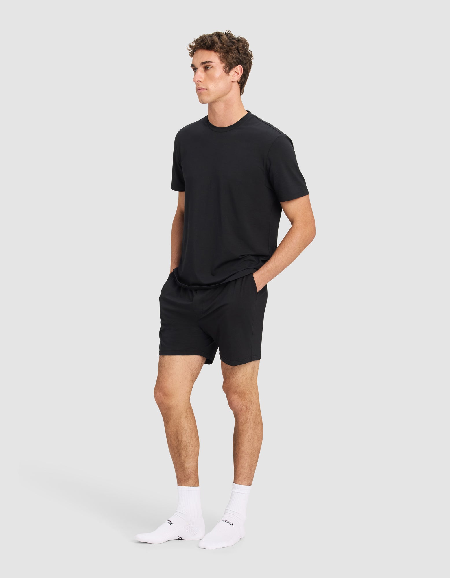 A young man stands against a plain light gray background wearing the Cozy Earth Men's First Layer Pajama Short Sleeve Tee & Short Set, paired with white socks and sneakers, hands in his pockets and a neutral expression.