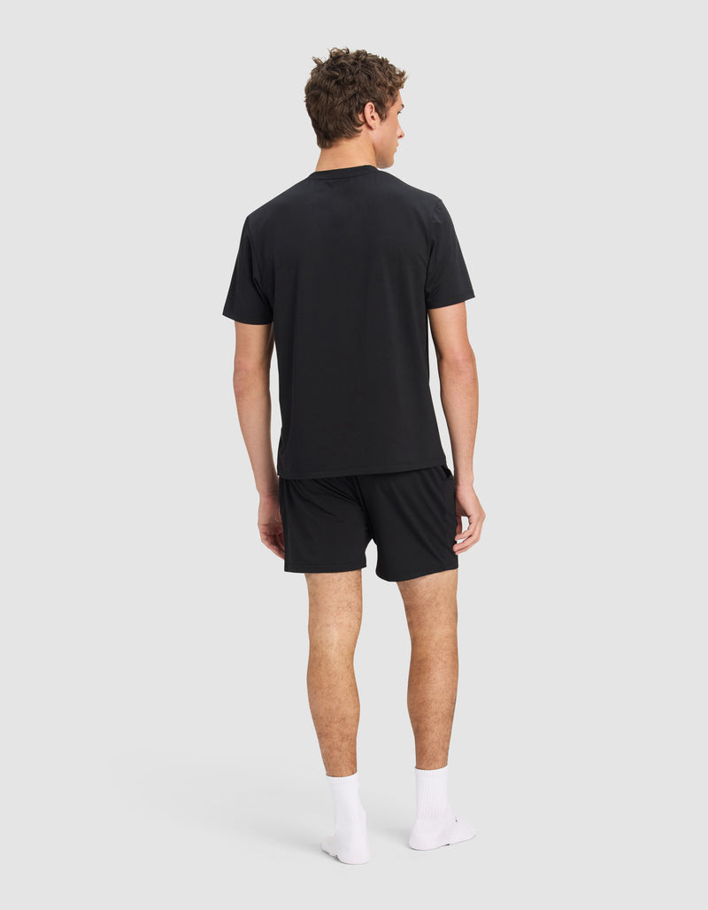 A man stands facing away, wearing the Cozy Earth Men's First Layer Pajama Short Sleeve Tee & Short Set in black, with white socks and no shoes, against a plain light gray background.