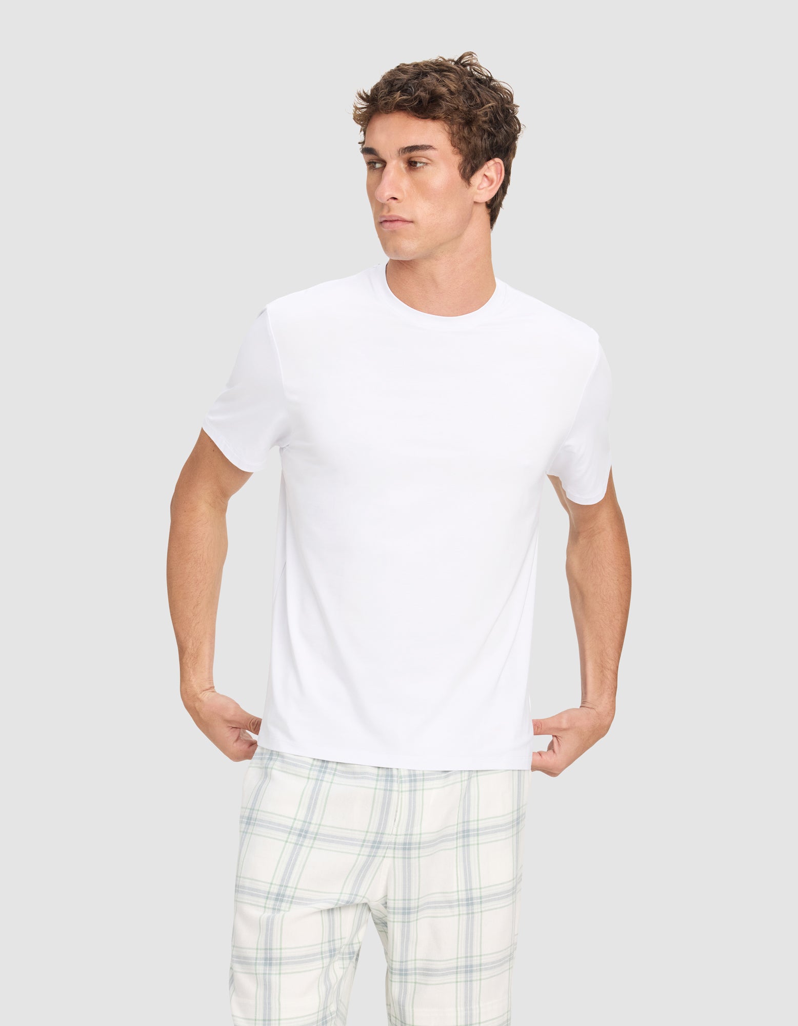 A young man models the Cozy Earth Men's First Layer Pajama Short Sleeve Tee and light plaid pajama pants against a light gray background, looking to his left while holding the hem of his shirt.