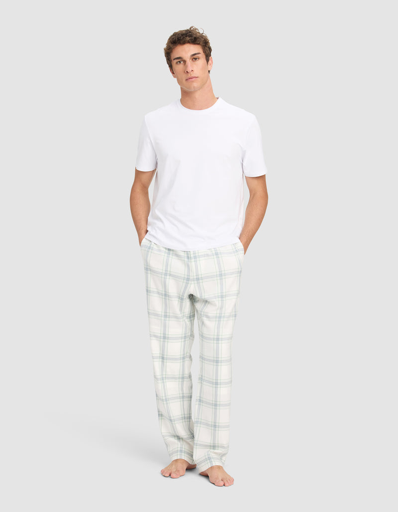 A young man stands barefoot against a plain background, wearing the Cozy Earth Men's First Layer Pajama Short Sleeve Tee and white plaid pajama pants, with his hands in his pockets.