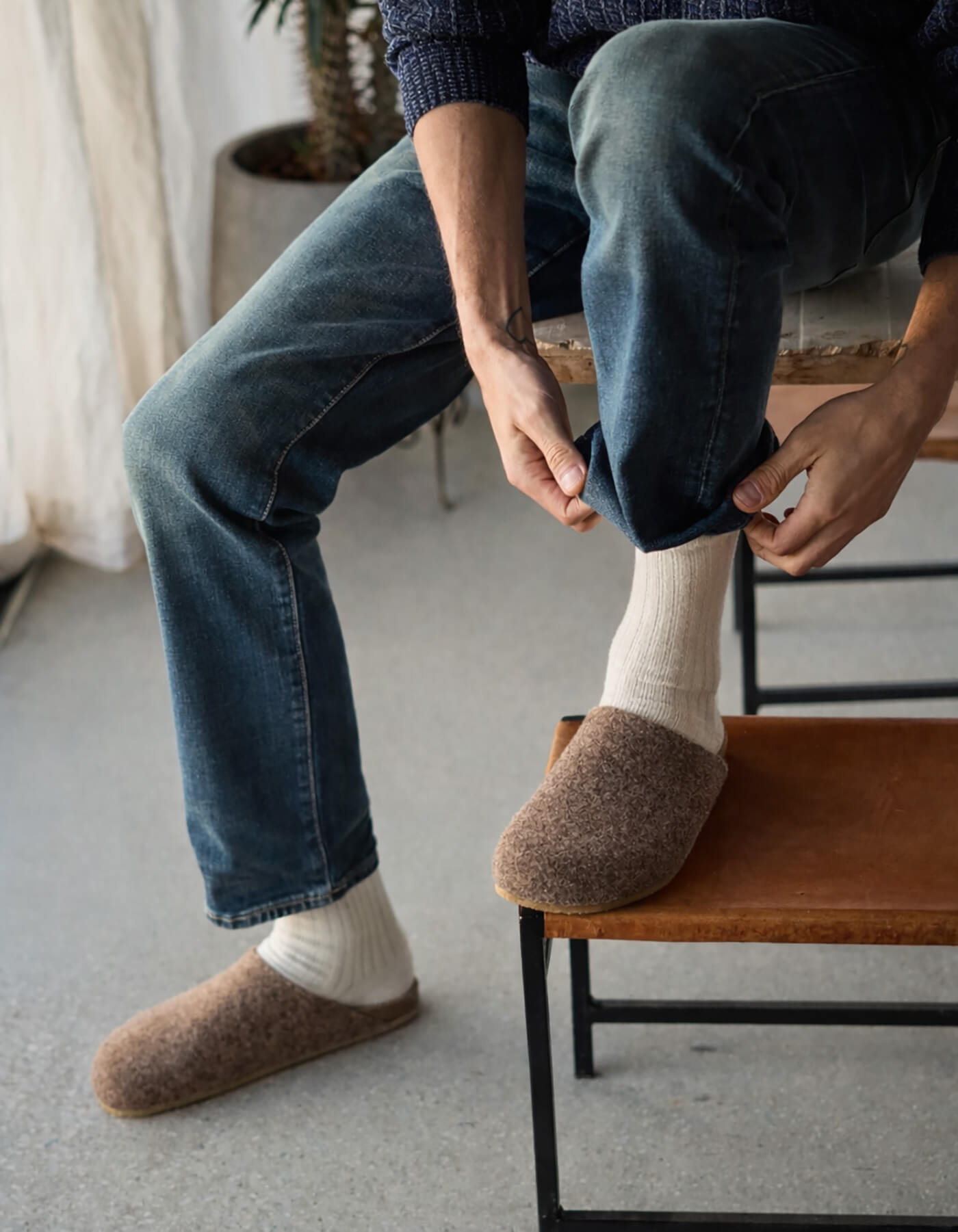 A person in a blue sweater and jeans sits on a chair, adjusting their pant leg. They're wearing white socks with brown Lakehouse Clogs by Cozy Earth. A plant and curtain are visible in the background. 