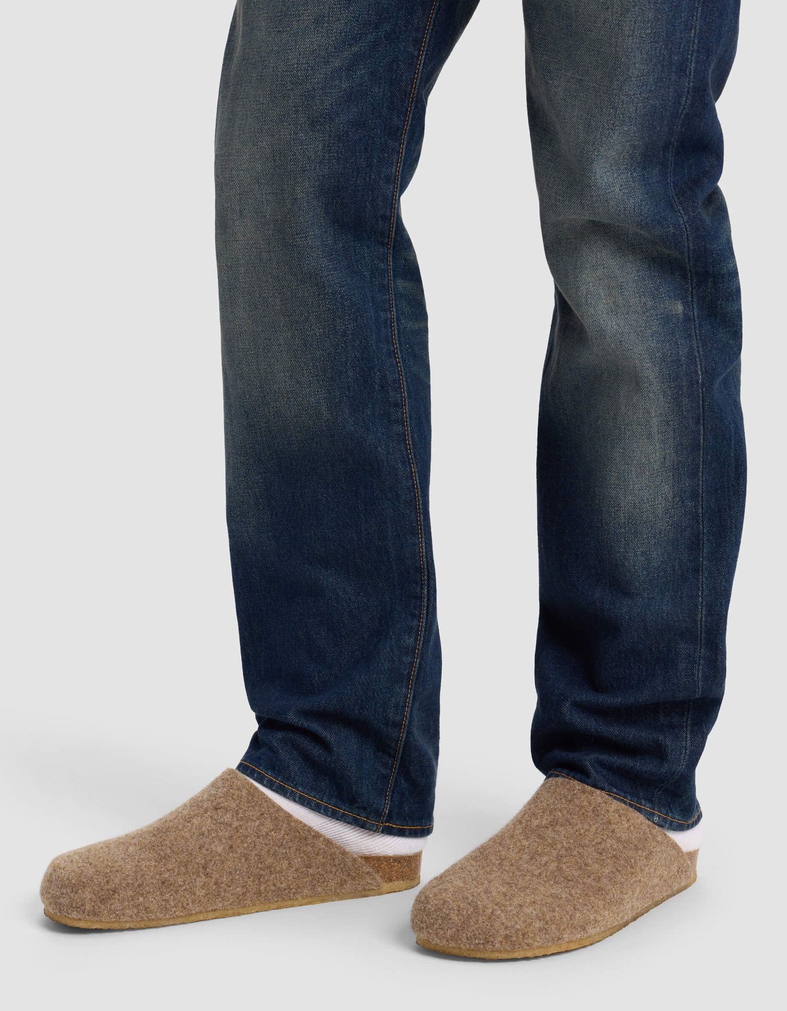 A person wearing blue jeans and Cozy Earth's Lakehouse Clog in tan stands on a light gray background, with only their lower legs and feet visible.