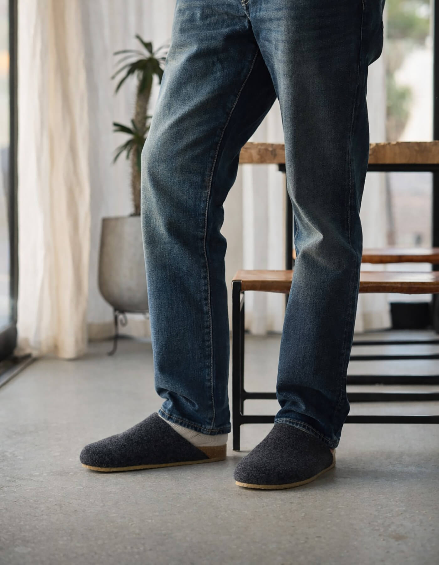 A person in blue jeans and Cozy Earth's graphite Lakehouse Clogs stands in a sunlit room with hands in pockets. A wooden bench, a potted plant, and sheer curtains are visible in the background. 