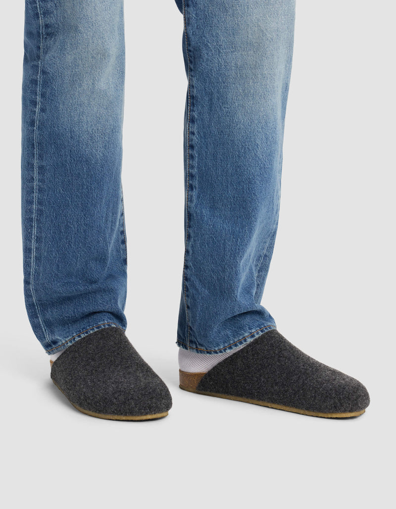 A person wears Cozy Earth's Lakehouse Clog slippers with blue jeans, standing on a light background. Only the lower legs and feet are visible.