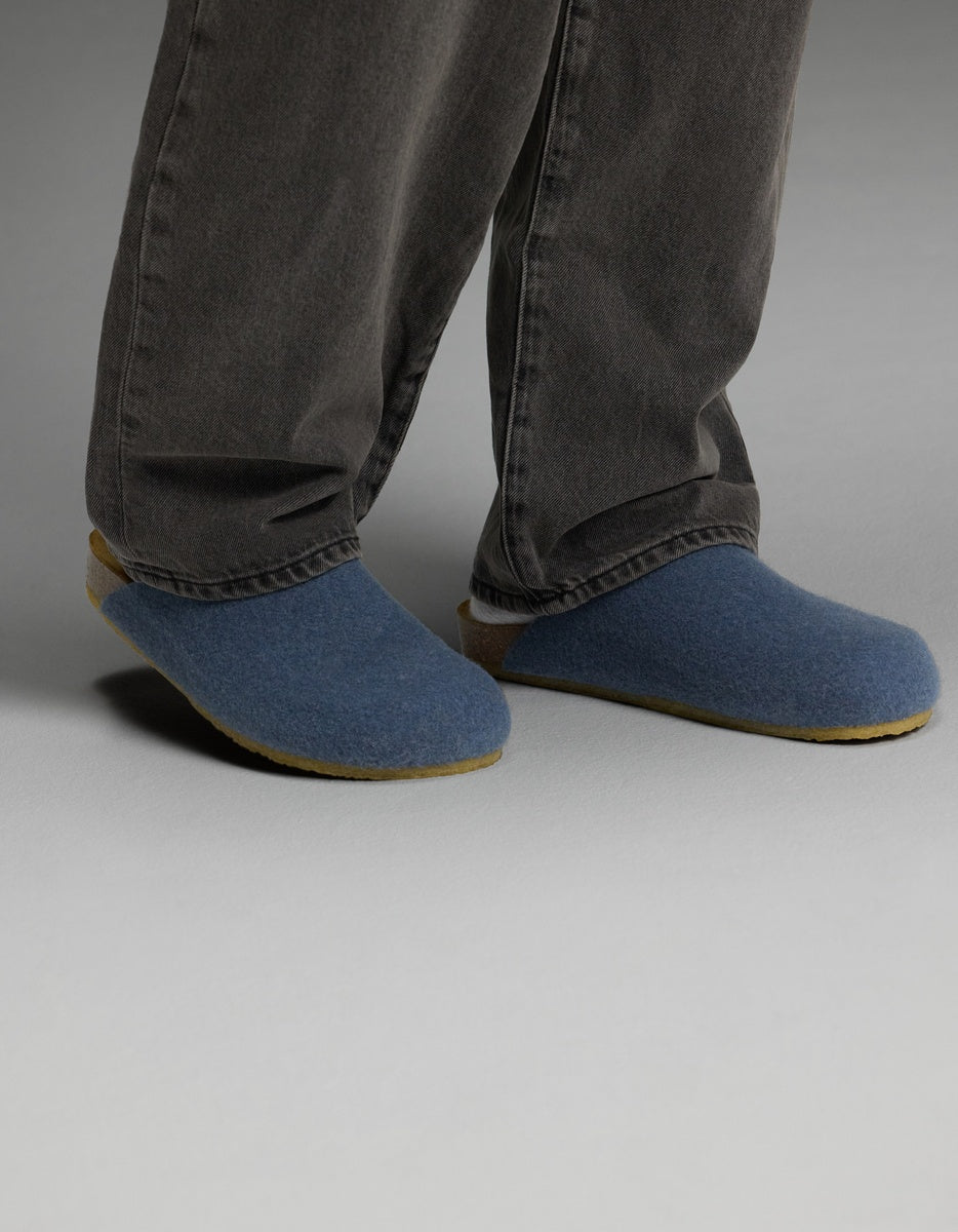A person wearing dark gray pants and Cozy Earth Lakehouse Clogs in Light Blue stands on a light gray floor, with only their lower legs and feet visible.