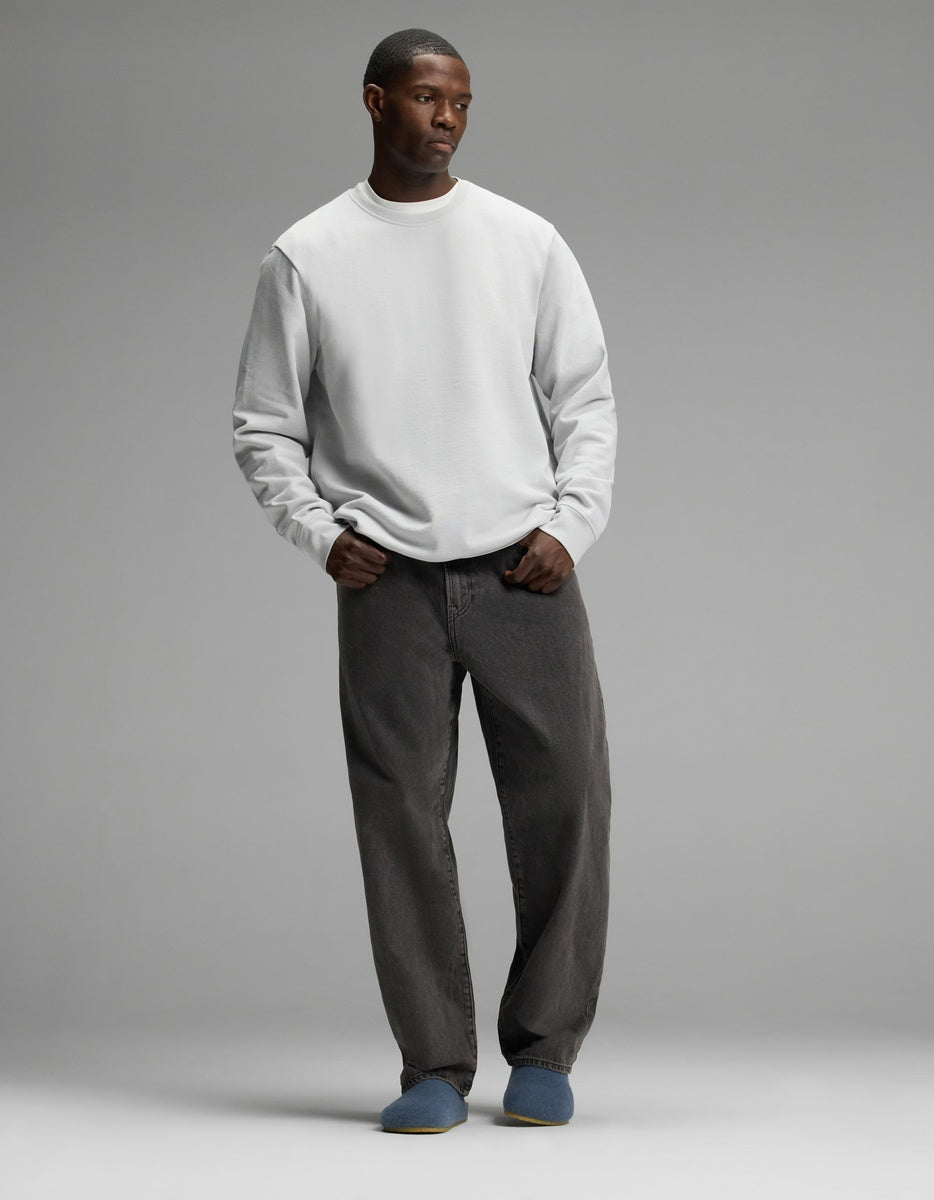 A man stands against a plain gray background, wearing a light gray sweatshirt, loose dark gray jeans, and Cozy Earth’s Lakehouse Clog slippers in blue. His hands are in his pockets and he has a neutral expression.