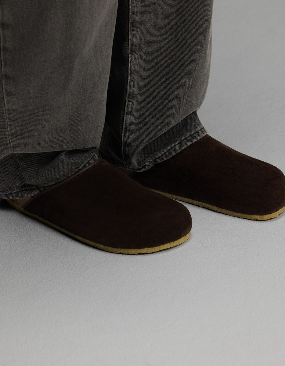 A person wearing the Cozy Earth Lakehouse Clog in Suede—dark brown slippers with light brown soles—paired with gray jeans stands on a light gray floor.|Color:Espresso