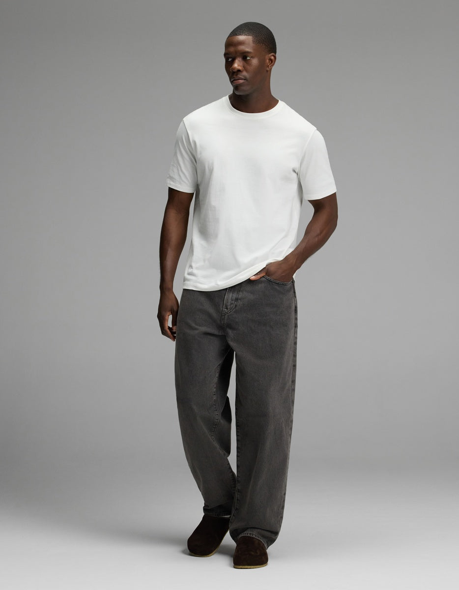 A man stands with one hand in his pocket against a plain gray background, wearing loose-fitting dark gray jeans, a plain white t-shirt, and Cozy Earth’s Lakehouse Clog in Suede.