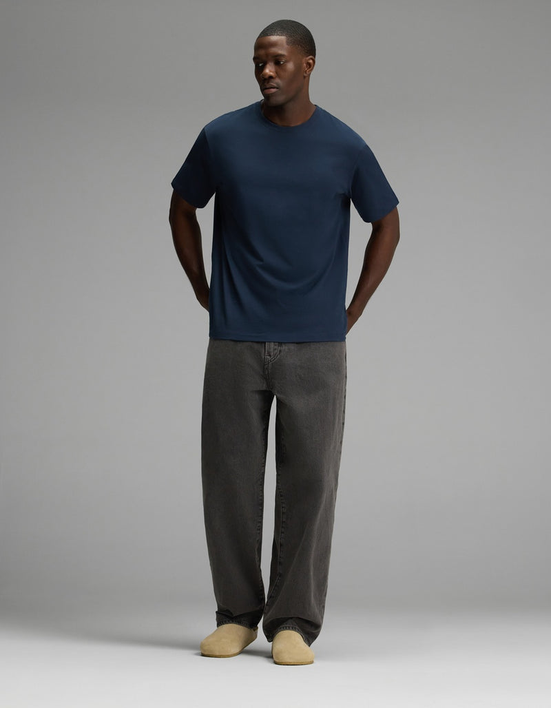 A person stands against a plain gray background, wearing a navy blue t-shirt, loose-fitting dark gray jeans, and the Lakehouse Clog in Suede by Cozy Earth, with their hands in their pockets.