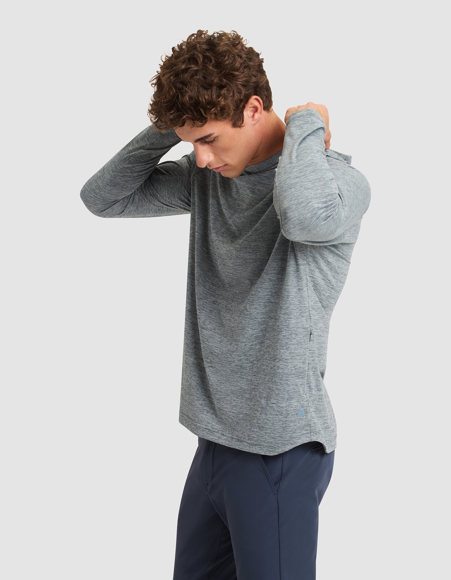 A man with curly brown hair wears the Cozy Earth Men's Lightweight Performance Hoodie in light gray and navy pants, adjusting his collar while looking down against a plain light background. 