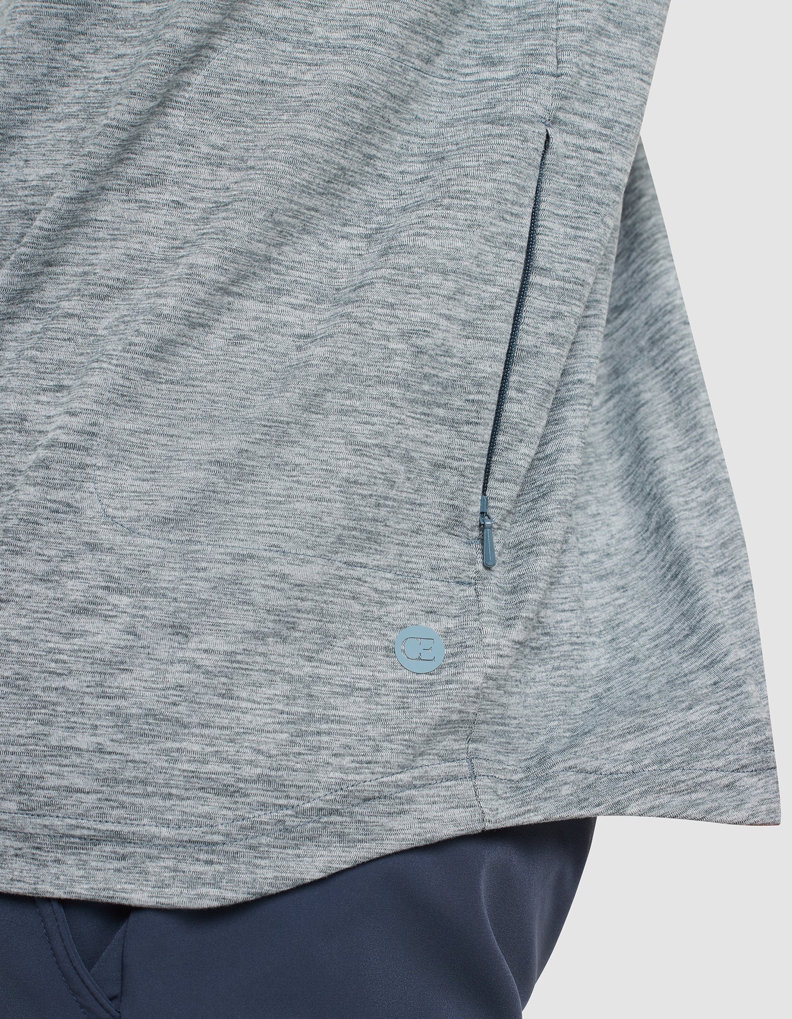 Close-up of a heather gray zippered pocket on the Cozy Earth Men's Lightweight Performance Hoodie, featuring a small round blue logo near the seam, paired with dark blue pants. 