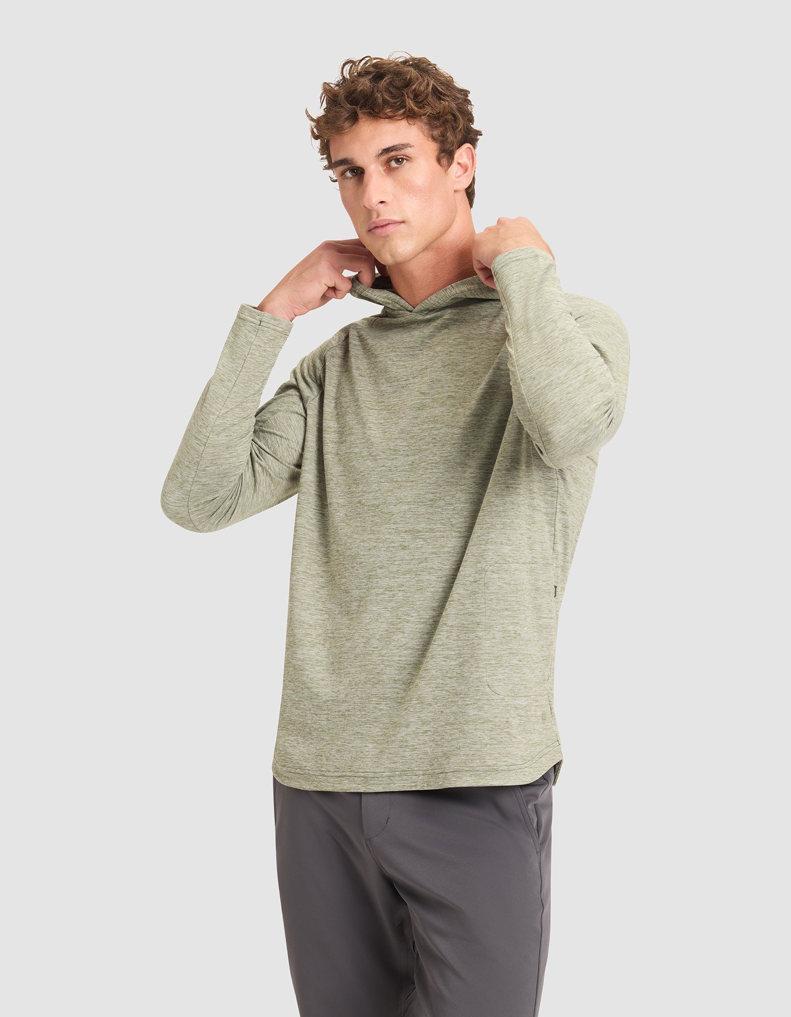 A young man adjusts the hood of a Cozy Earth Men's Lightweight Performance Hoodie, standing against a plain light gray background while wearing gray pants. |Color:Heather Olive