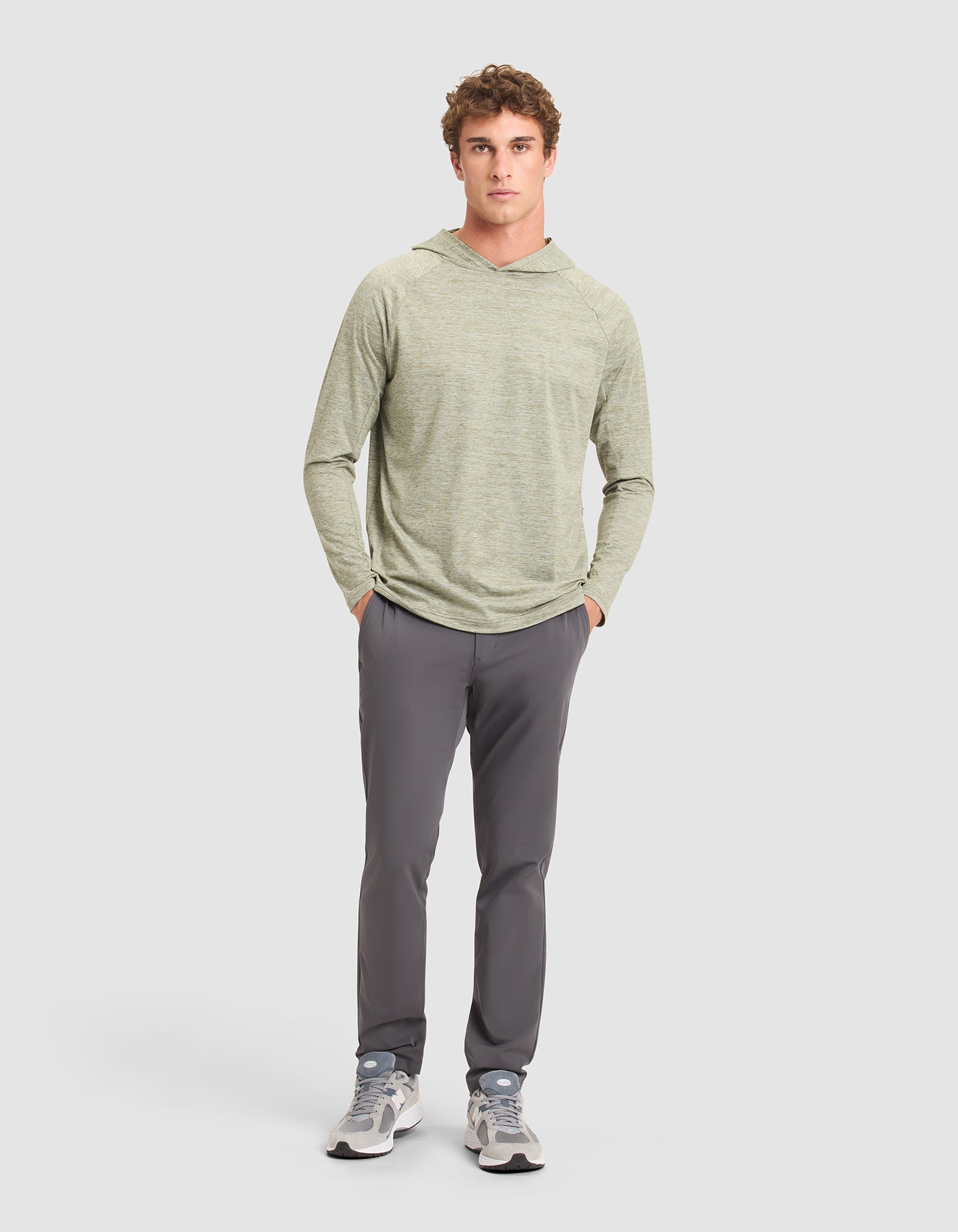 A young man stands against a plain background wearing the Cozy Earth Men's Lightweight Performance Hoodie in light green, paired with gray pants and gray sneakers, with his hands in his pockets. |Color:Heather Olive