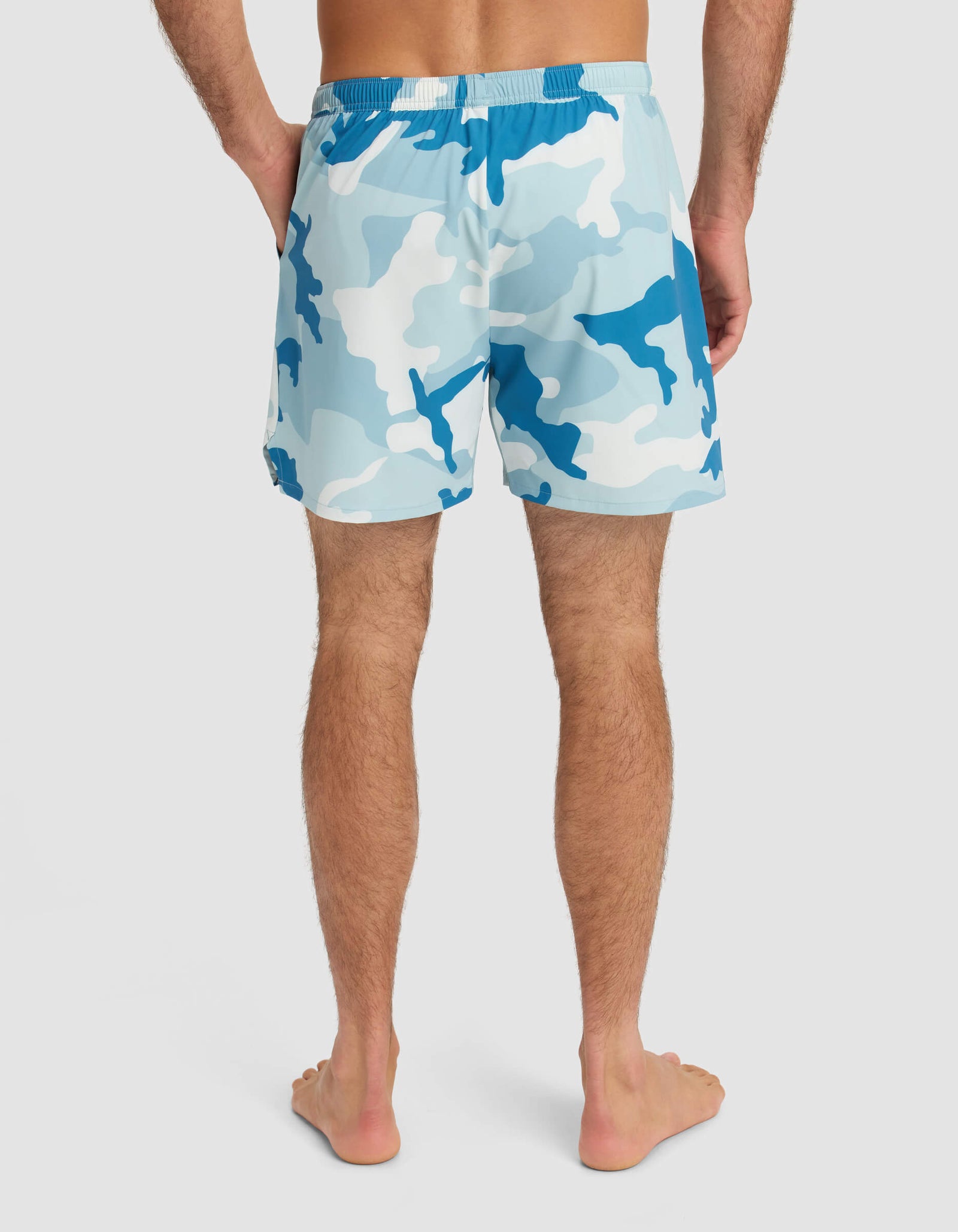 A person stands barefoot, facing away from the camera, wearing Cozy Earth Men's Day & Night Short 5" in blue and white camo. The background is plain and light-colored.