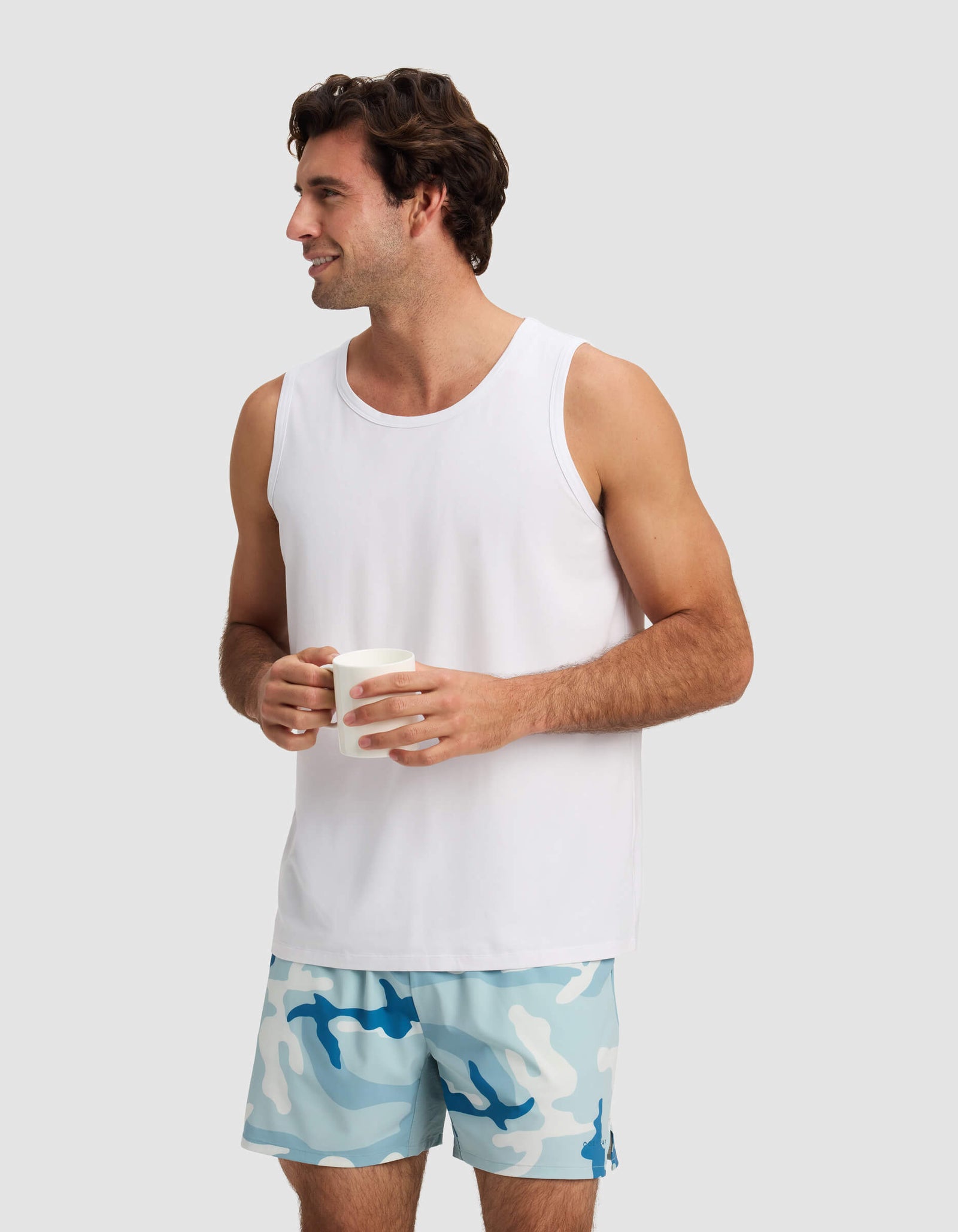 A man wearing Cozy Earth's Men's Day & Night Short 5" and a white tank top smiles while holding a white mug, standing against a plain light gray background.