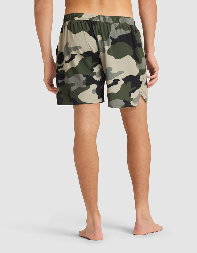 A person stands barefoot, facing away, wearing Cozy Earth Men's Day & Night Shorts 5" with an elastic waistband and a green, beige, and black camouflage pattern against a plain light background.