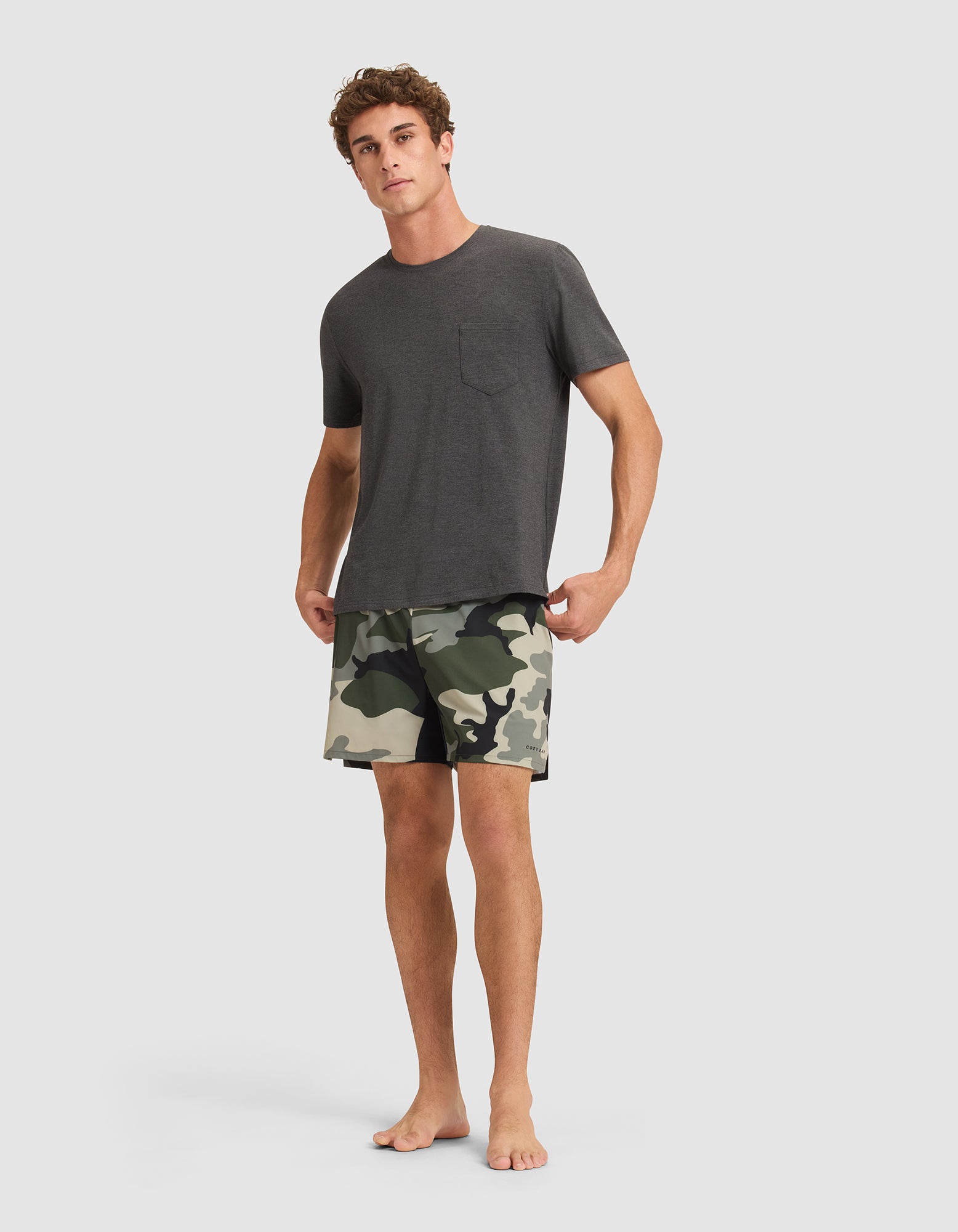 A man stands barefoot against a plain background, wearing a dark gray short-sleeve t-shirt with a chest pocket and Cozy Earth Men's Day & Night Shorts 5" in green camouflage. 
