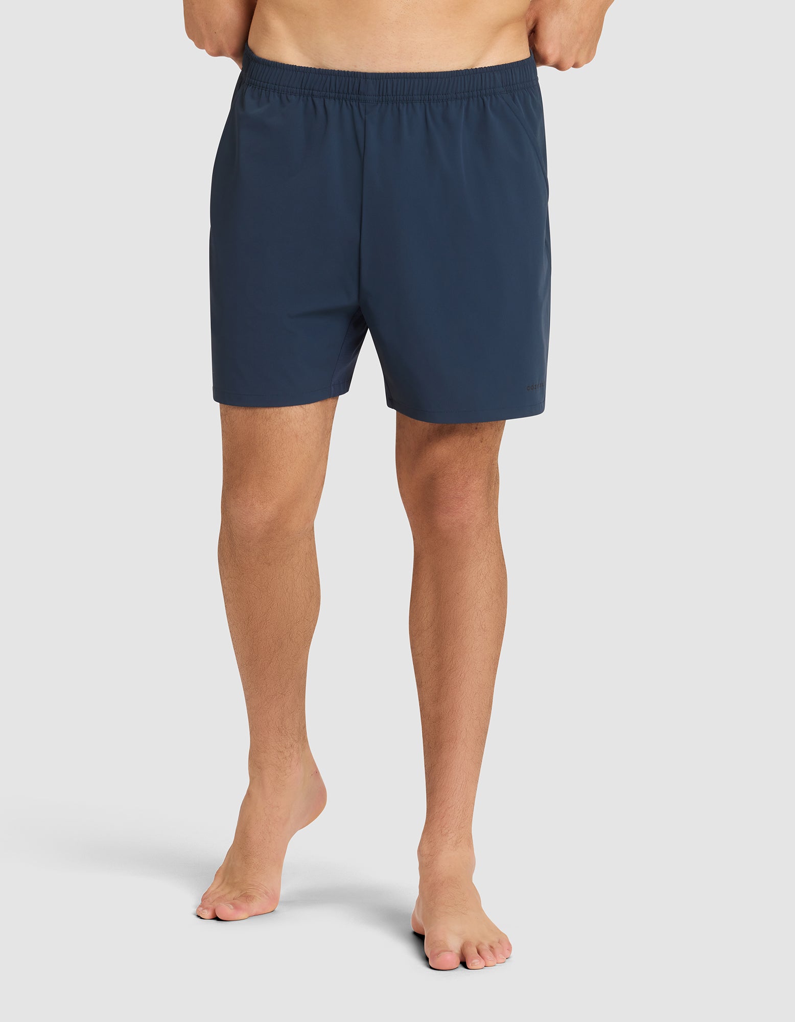 A person stands barefoot against a plain light background, wearing Cozy Earth Men's Day & Night Shorts 5" in dark blue with an elastic waistband. The upper body and head are not visible. 