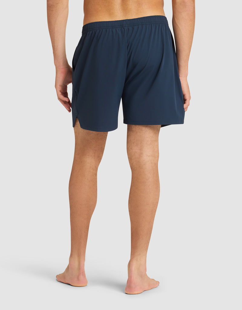 A person stands barefoot facing away, wearing Cozy Earth Men's Performance Sleep Shorts in navy with an elastic waistband and side slits, set against a plain light background.