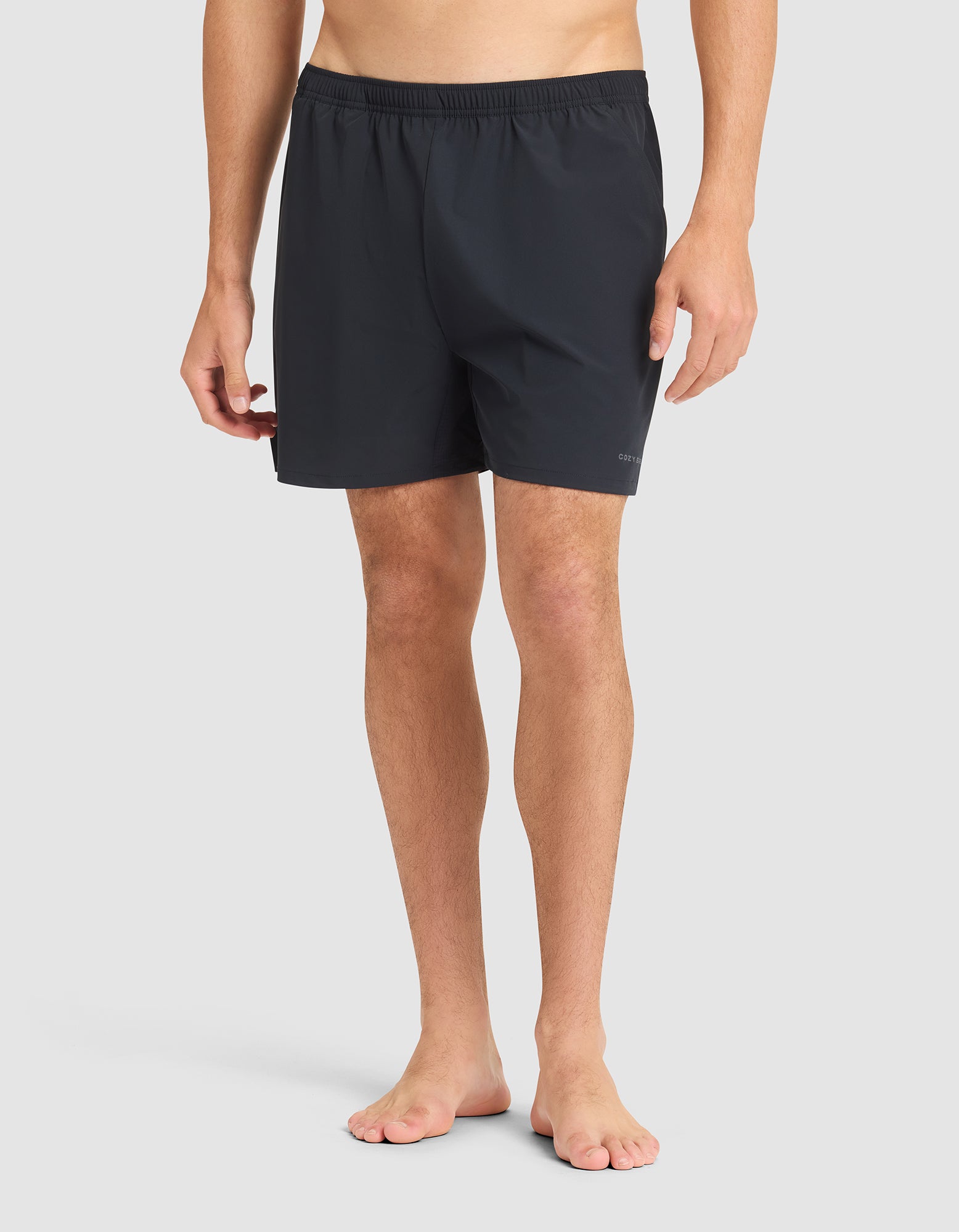 A man stands barefoot against a plain background, wearing Cozy Earth Men's Day & Night Shorts 5" with an elastic waistband. His upper body is not visible in the image. 