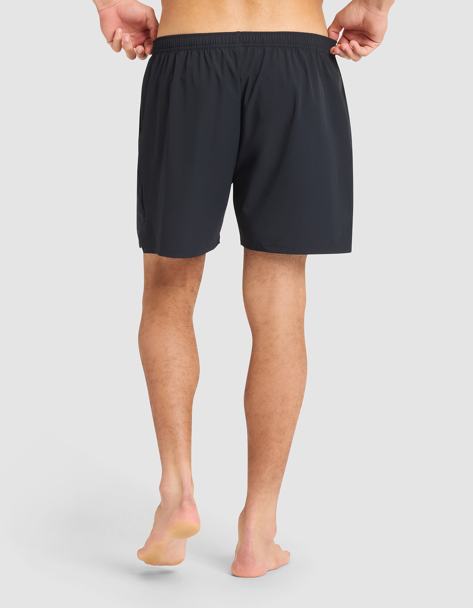 A barefoot man faces away from the camera, holding the waistband of Cozy Earth Men's Day & Night Shorts 5" in black against a plain light background. 