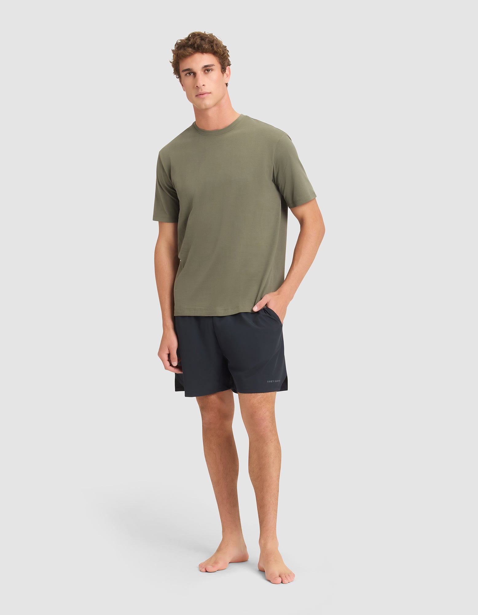 A young man stands barefoot against a plain background, wearing Cozy Earth Men's Day & Night Shorts 5" in black and an olive green short-sleeve t-shirt. His left hand is in his pocket, and he has a neutral expression. 