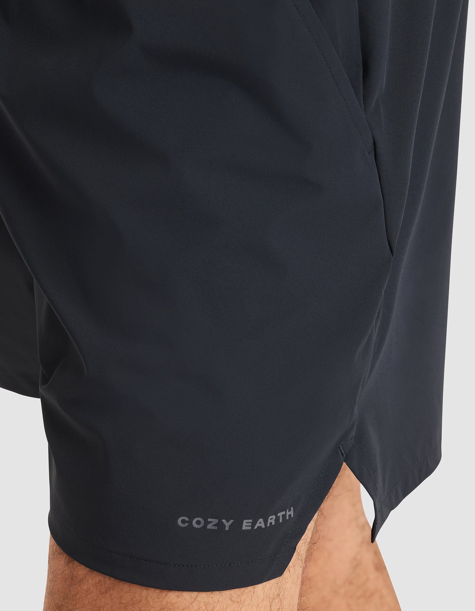 Close-up of a person wearing Cozy Earth Men's Day & Night Shorts 5" in black, with "COZY EARTH" printed in white near the bottom hem, standing against a plain light background. 