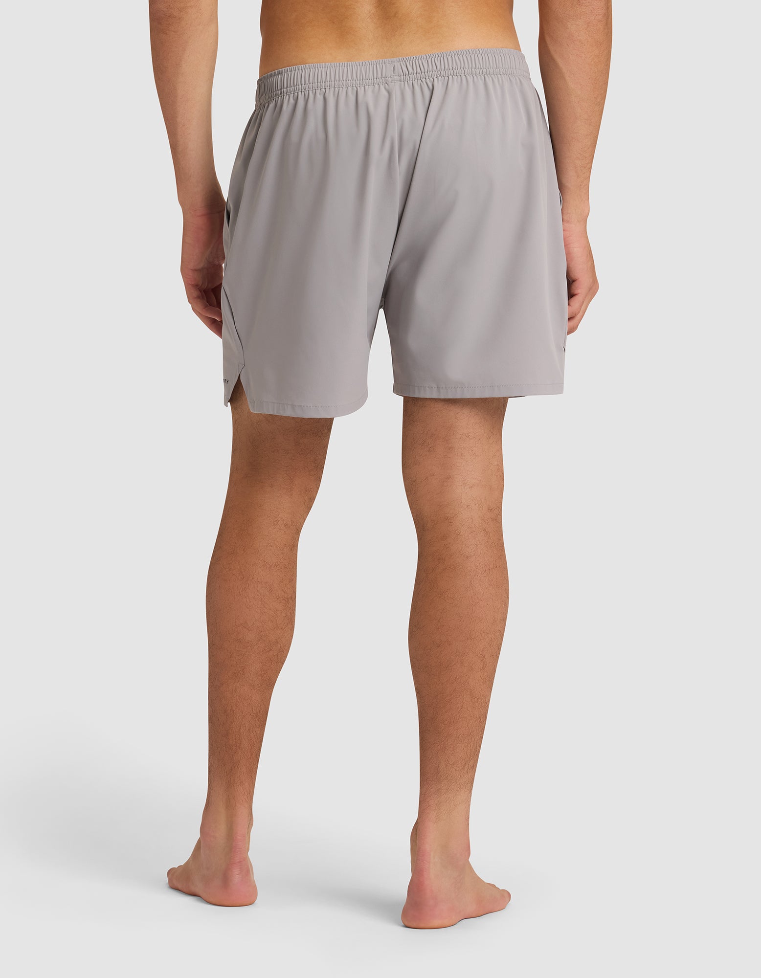 A man stands barefoot, facing away from the camera, wearing Cozy Earth Men's Day & Night Shorts 5" in light gray with an elastic waistband, set against a plain white background. 