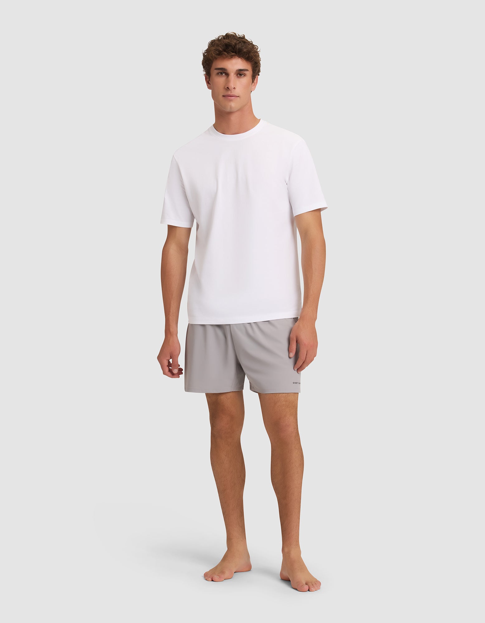 A young man stands barefoot against a light grey background, facing forward with a neutral expression. He is wearing Cozy Earth Men's Day & Night Shorts 5" in light grey and a plain white short-sleeve t-shirt. 
