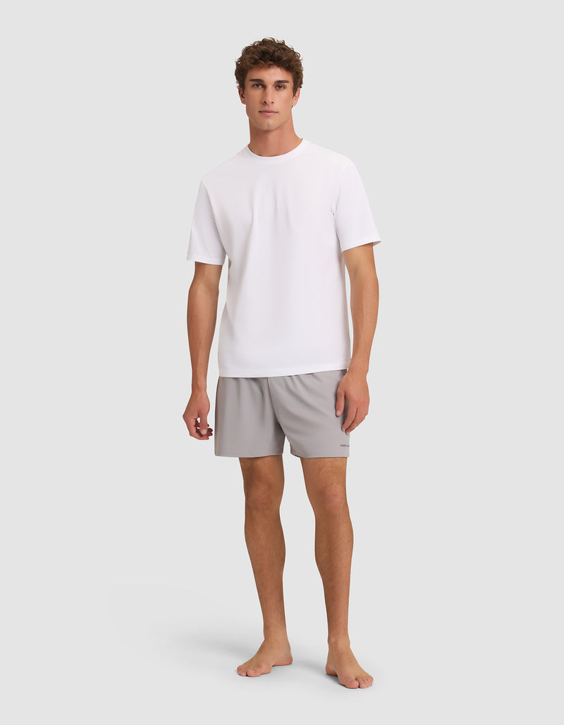 A young man stands barefoot against a light grey background, facing forward with a neutral expression. He is wearing Cozy Earth's Men's Performance Sleep Short in light grey and a plain white short-sleeve t-shirt.