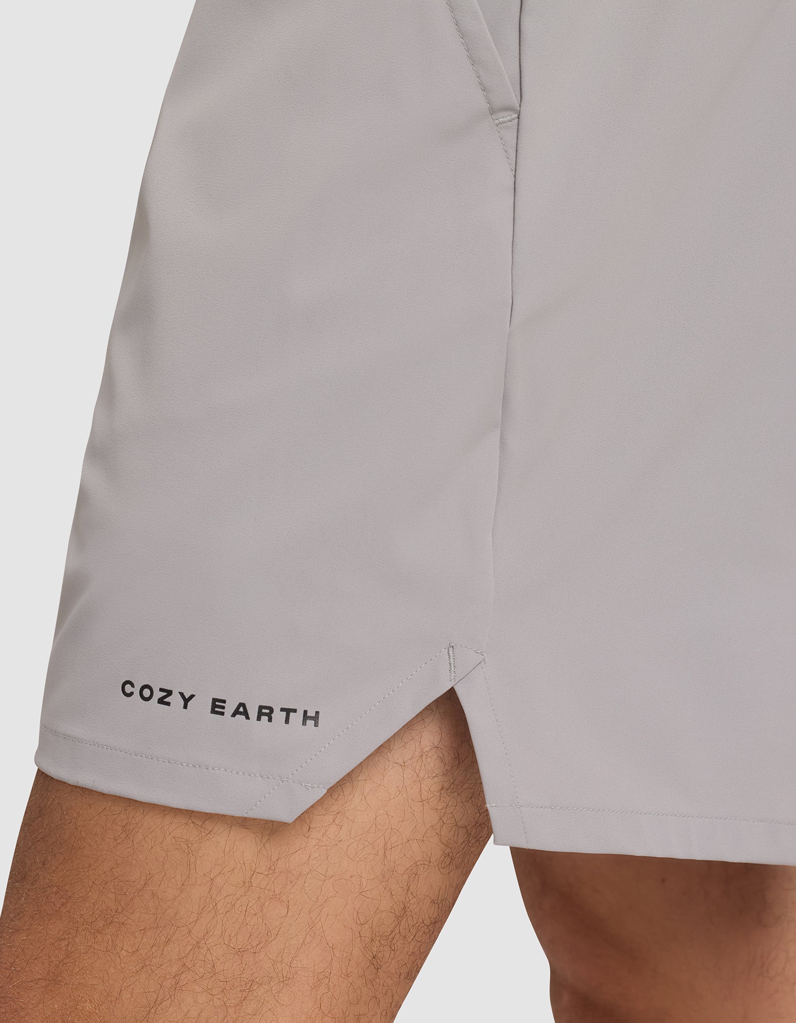 A close-up of a person wearing Cozy Earth Men's Performance Sleep Shorts in light gray, featuring the brand name near the left hem, a small side slit, and a visible pocket seam. |Color:Stone