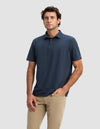 A man wears the Cozy Earth Men's Pique Polo in navy blue and beige pants, standing against a plain light gray background with one hand in his pocket and a neutral expression.|Color:Eclipse