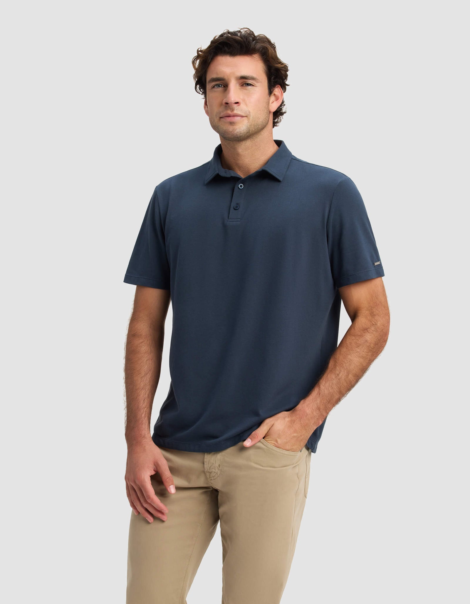 A man wears the Cozy Earth Men's Pique Polo in navy blue and beige pants, standing against a plain light gray background with one hand in his pocket and a neutral expression.
