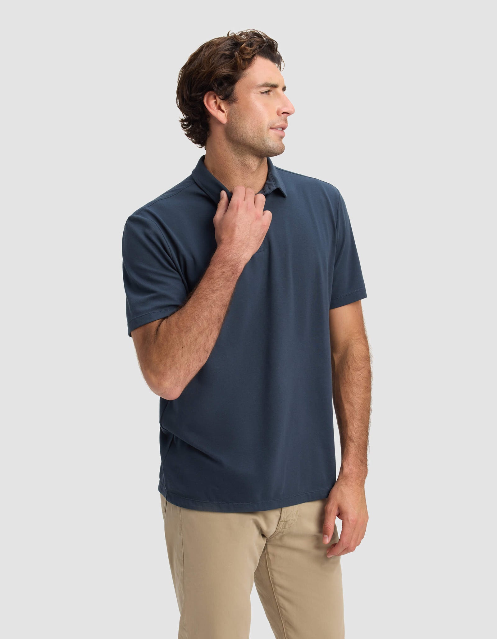 A man wearing the Cozy Earth Men's Pique Polo in navy blue and beige pants stands against a light gray background, looking to the side with one hand touching his collar.