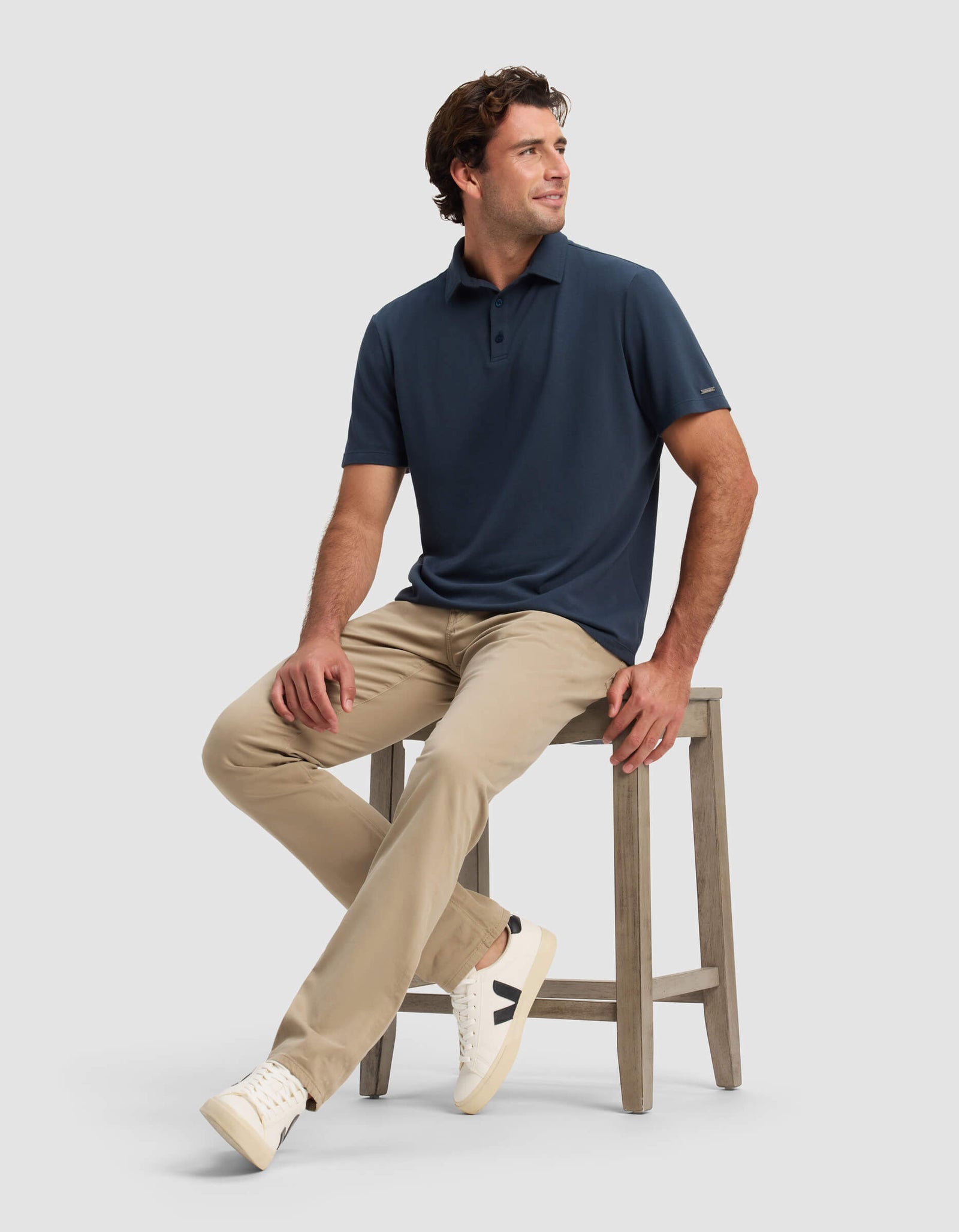 A man sits on a wooden stool against a plain light gray background, wearing Cozy Earth's Men's Pique Polo in navy, beige pants, and white sneakers with black details.