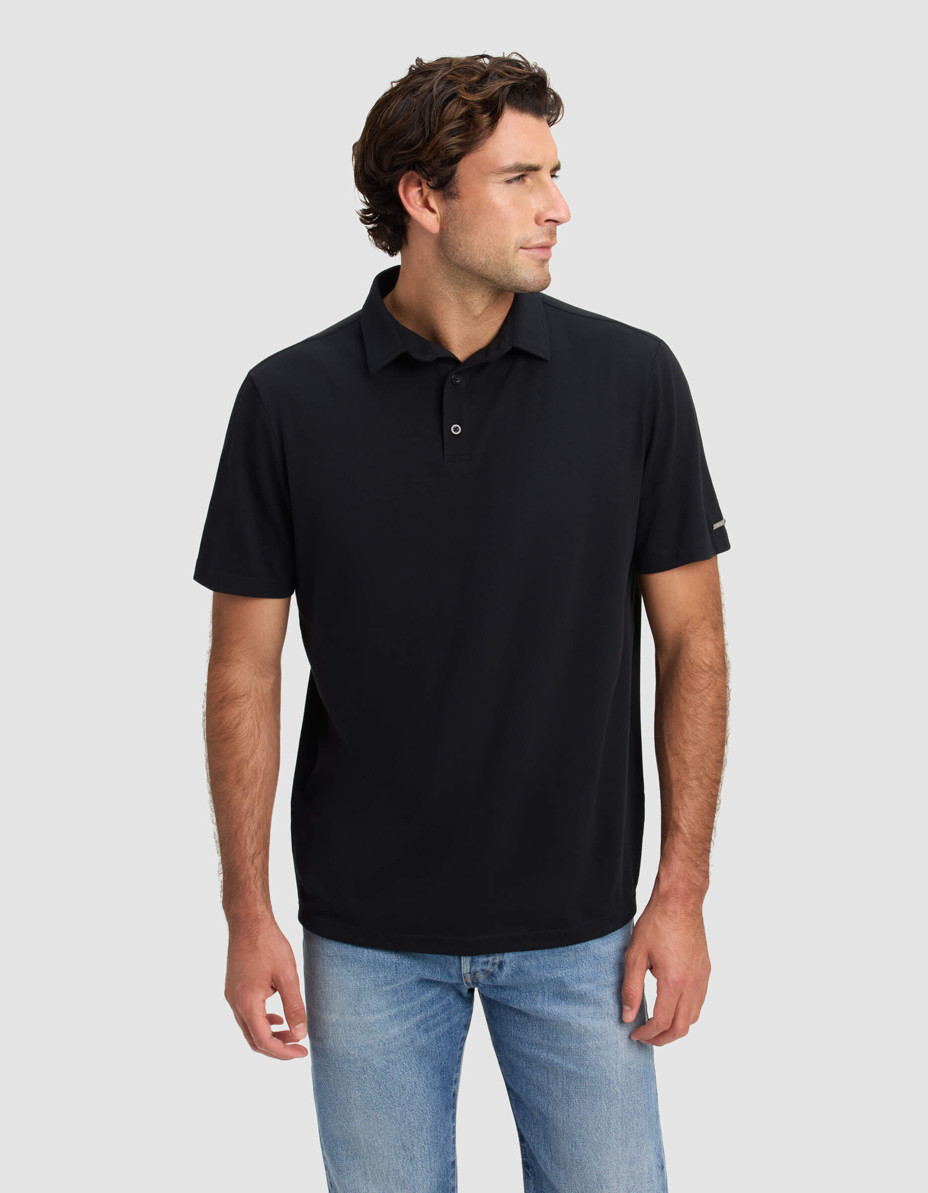 A man with wavy brown hair wears the Cozy Earth Men's Pique Polo in black and light blue jeans, standing against a plain light gray background and looking to his left.