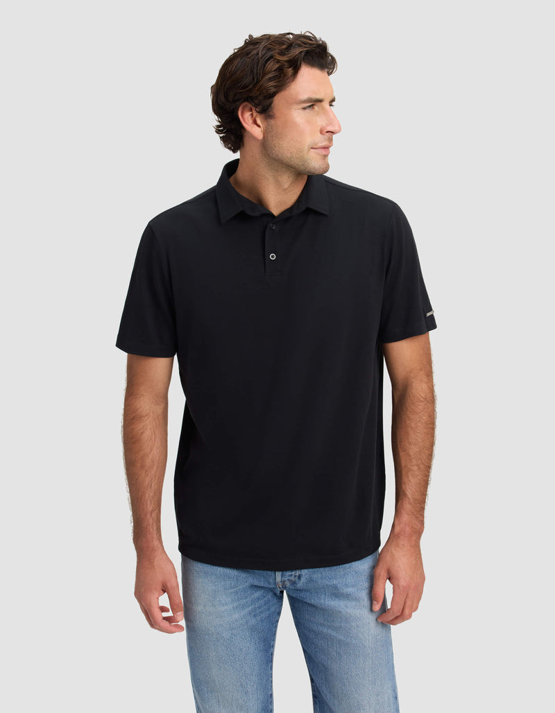 A man with wavy brown hair wears the Cozy Earth Men's Pique Polo in black and light blue jeans, standing against a plain light gray background and looking to his left.