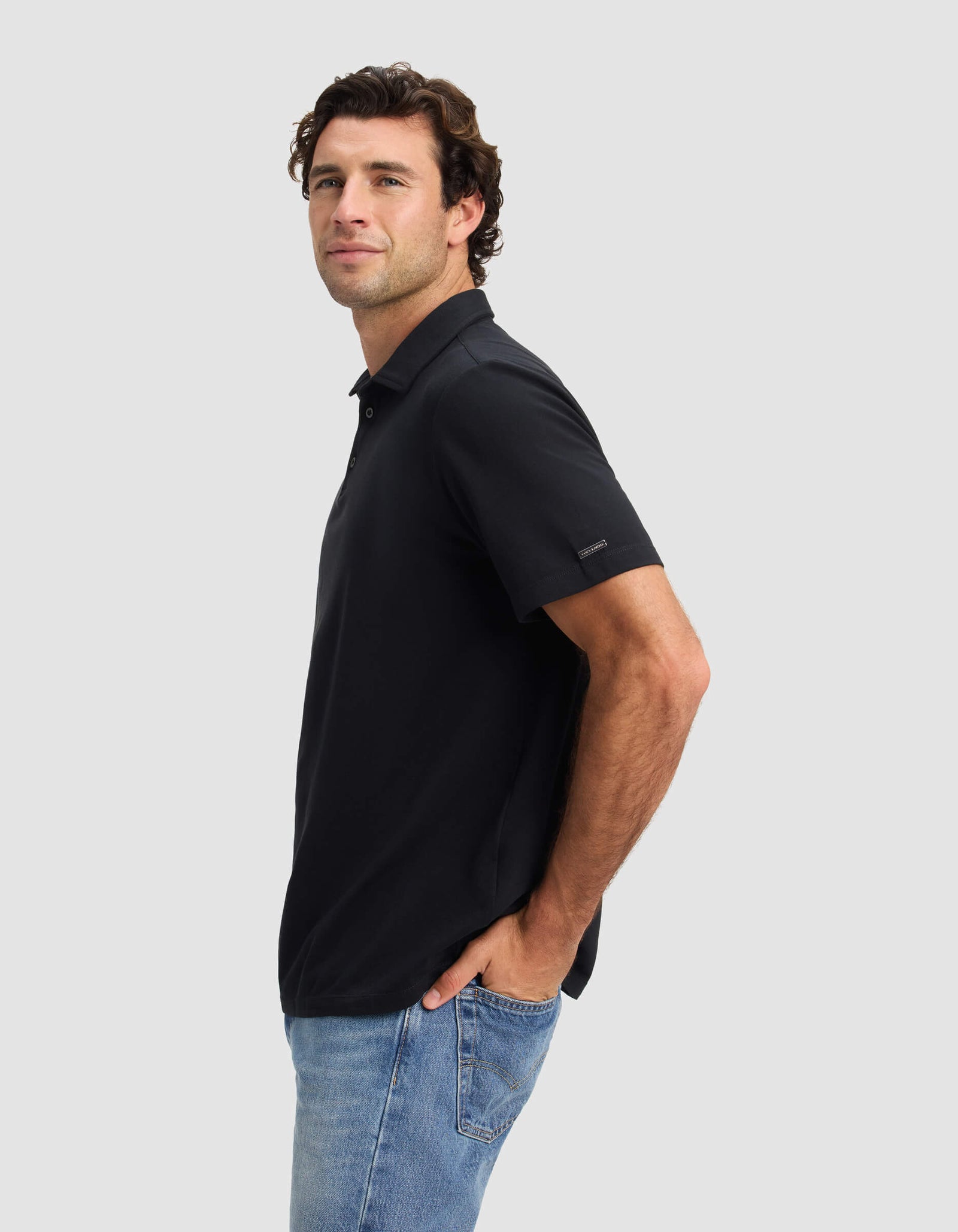 A man with wavy brown hair models the Cozy Earth Men's Pique Polo in black, paired with light blue jeans, standing sideways with hands in pockets and facing the camera against a plain light gray background.