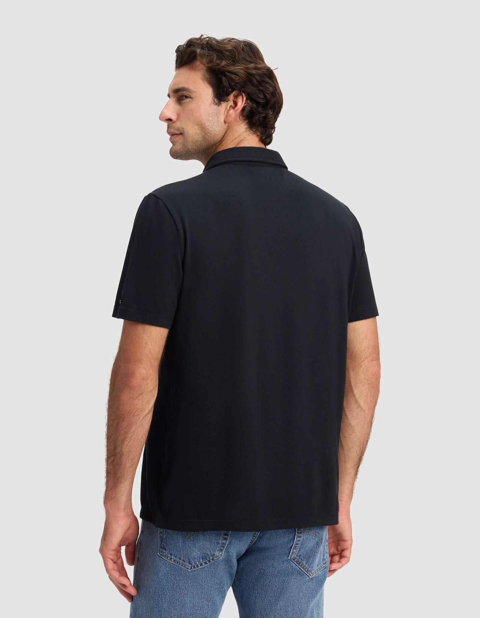 A man faces away wearing the Cozy Earth Men's Pique Polo in black with blue jeans against a light gray background, looking over his left shoulder with a neutral expression.