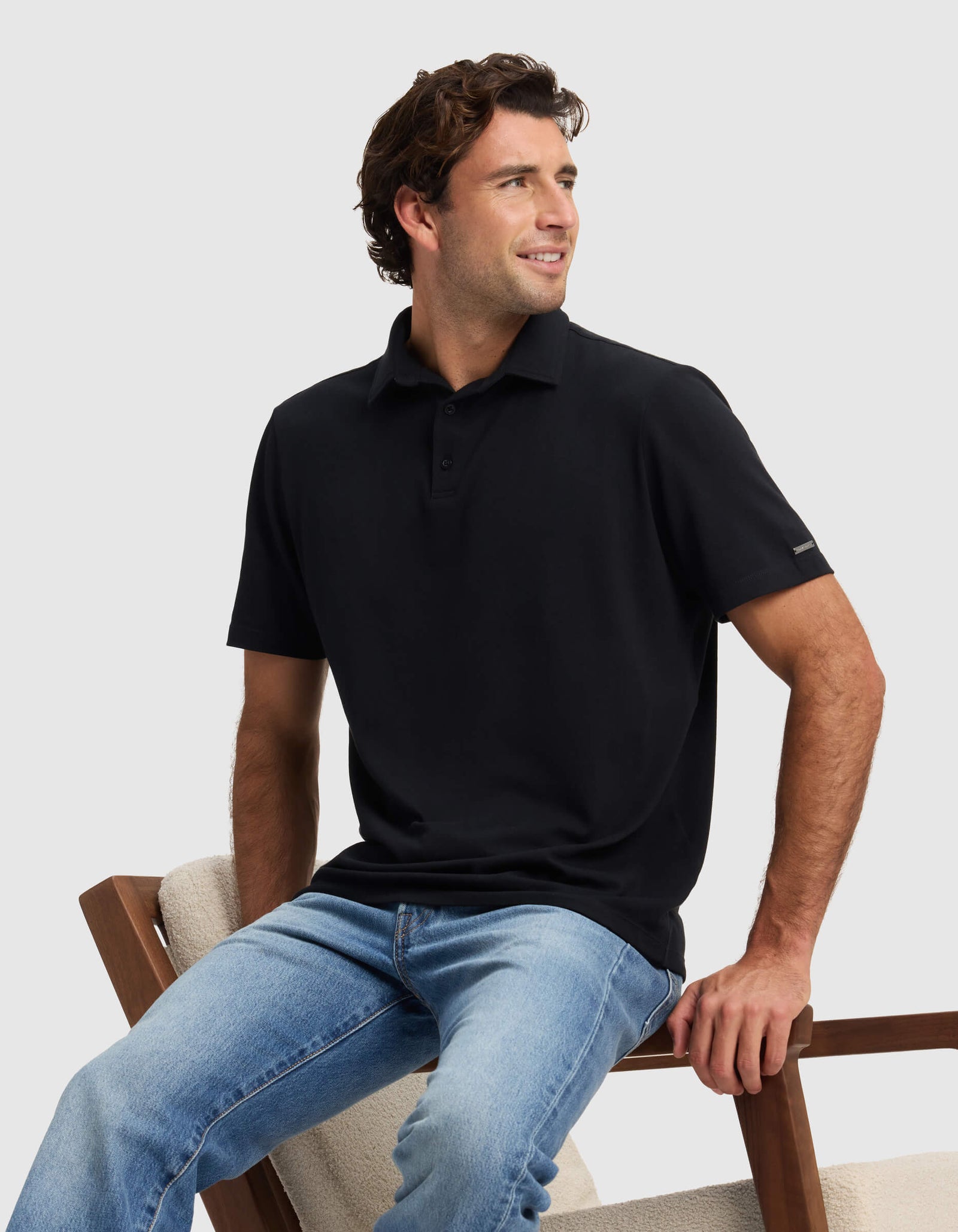 A man wears the Cozy Earth Men's Pique Polo in black and light blue jeans, sitting on a cushioned wooden chair and smiling to his right against a plain light gray background.