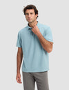 A man wearing the Cozy Earth Men's Pique Polo in light blue and gray pants stands against a plain light gray background, looking forward with one hand touching his collar.|Color:River