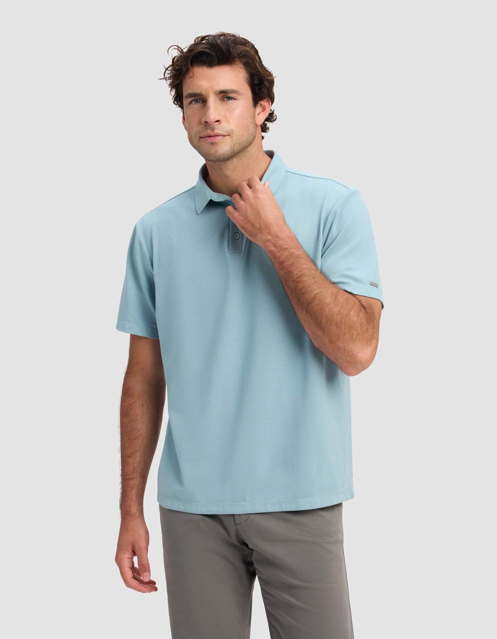 A man wearing the Cozy Earth Men's Pique Polo in light blue and gray pants stands against a plain light gray background, looking forward with one hand touching his collar.