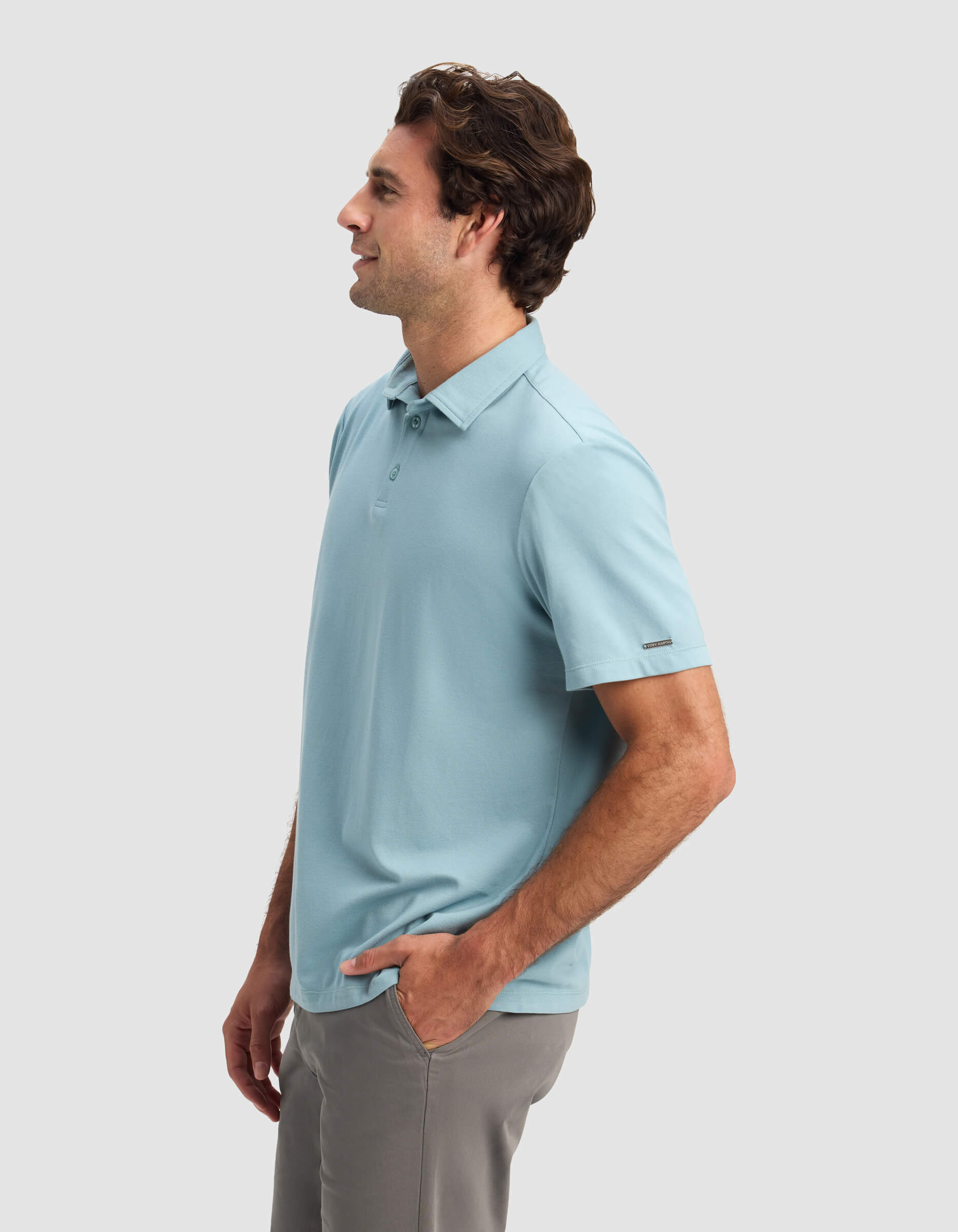 A man models the Cozy Earth Men's Pique Polo in light blue, paired with gray pants. He stands in profile with one hand in his pocket, facing left against a plain light gray background.|Color:River